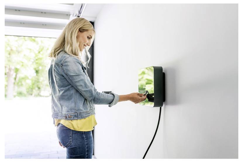 Frau, die ein Elektrofahrzeug an eine an der Wand montierte Ladestation in Innenräumen anschließt.