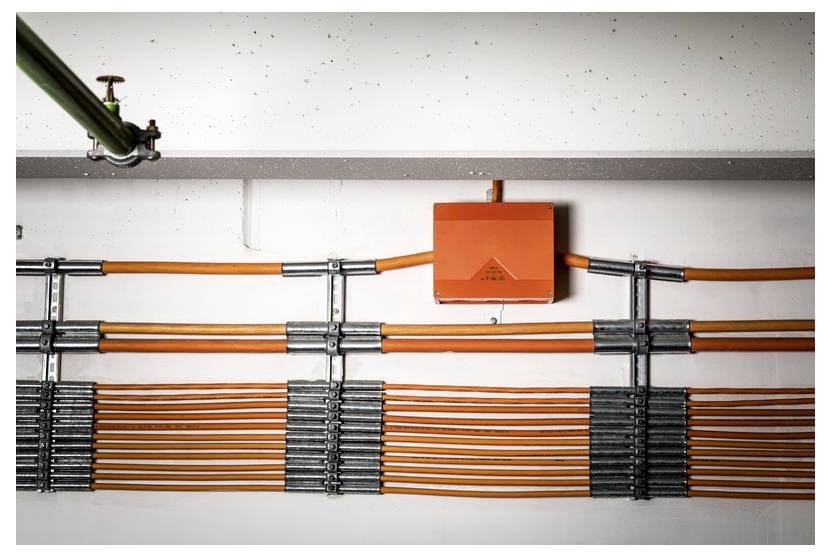 Orange Elektroverteilungskasten mit mehreren orangefarbenen Kabeln und metallischem Elektrorohr an einer Wand unter einem Deckenrohr montiert.