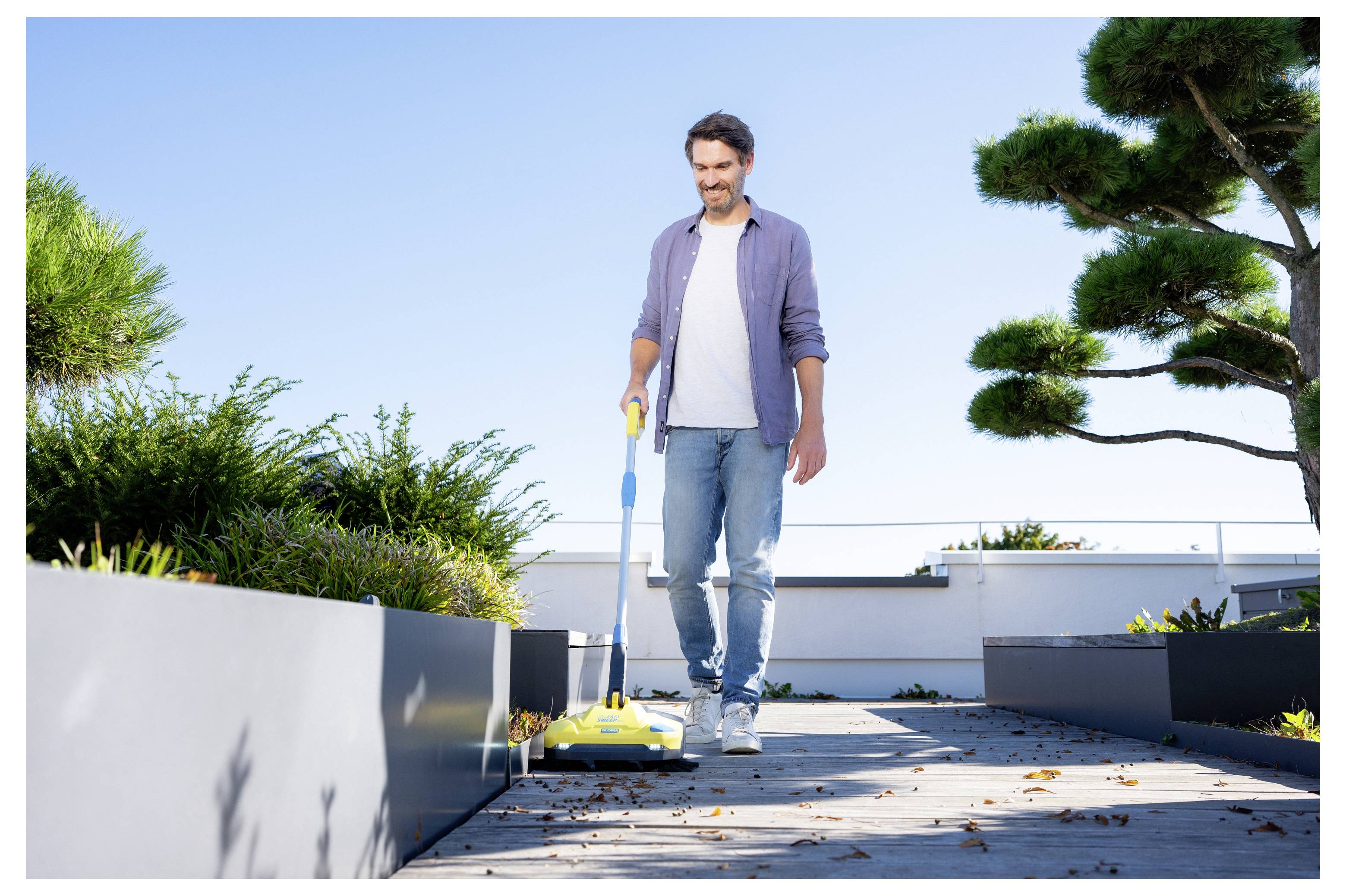 Ein Mann reinigt einen Dachgartenwegmit einem Elektrobesen, umgeben von grünen Pflanzen und klarem blauem Himmel, was auf Außenreinigung hindeutet.