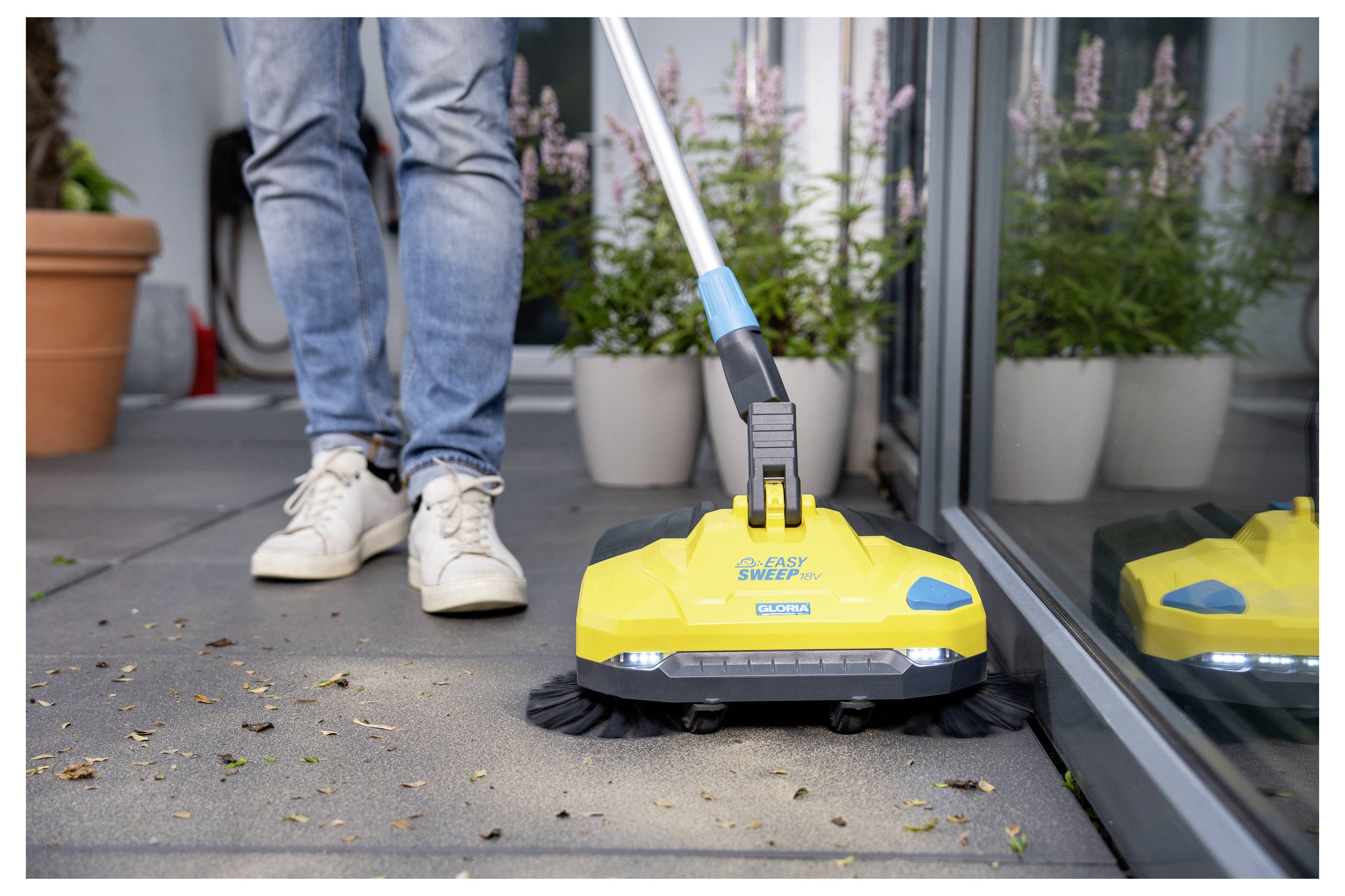 Person, die einen gelben Bodenkehrbesen auf einer Terrasse benutzt, mit Pflanzen im Hintergrund. Das Werkzeug ist mit Easy Sweep gekennzeichnet.