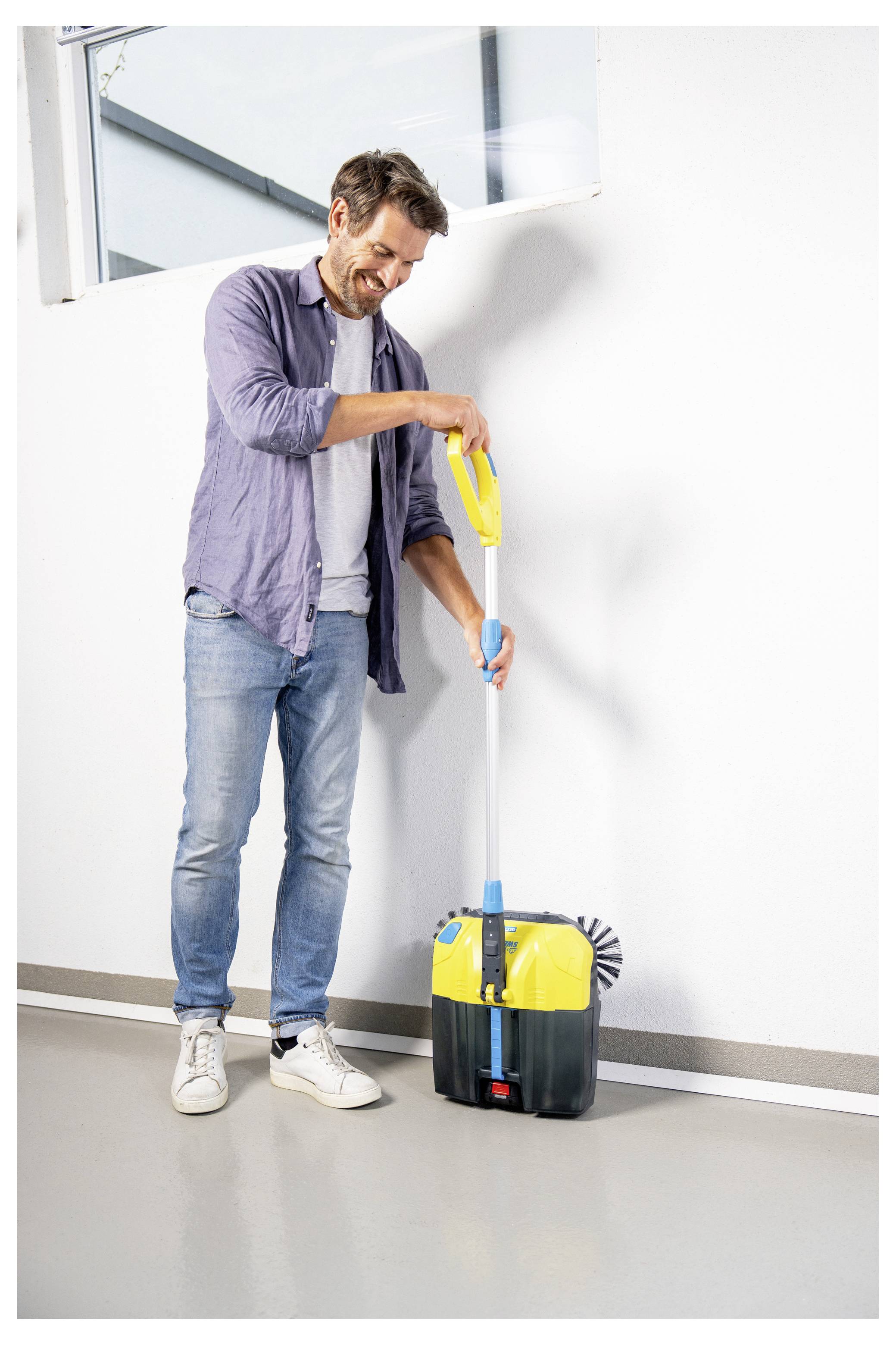 Eine Person, die lächelt, während sie ein gelb-schwarzes Reinigungswerkzeug mit langem Griff und Bürste gegen eine weiße Wand im Innenraum verwendet.