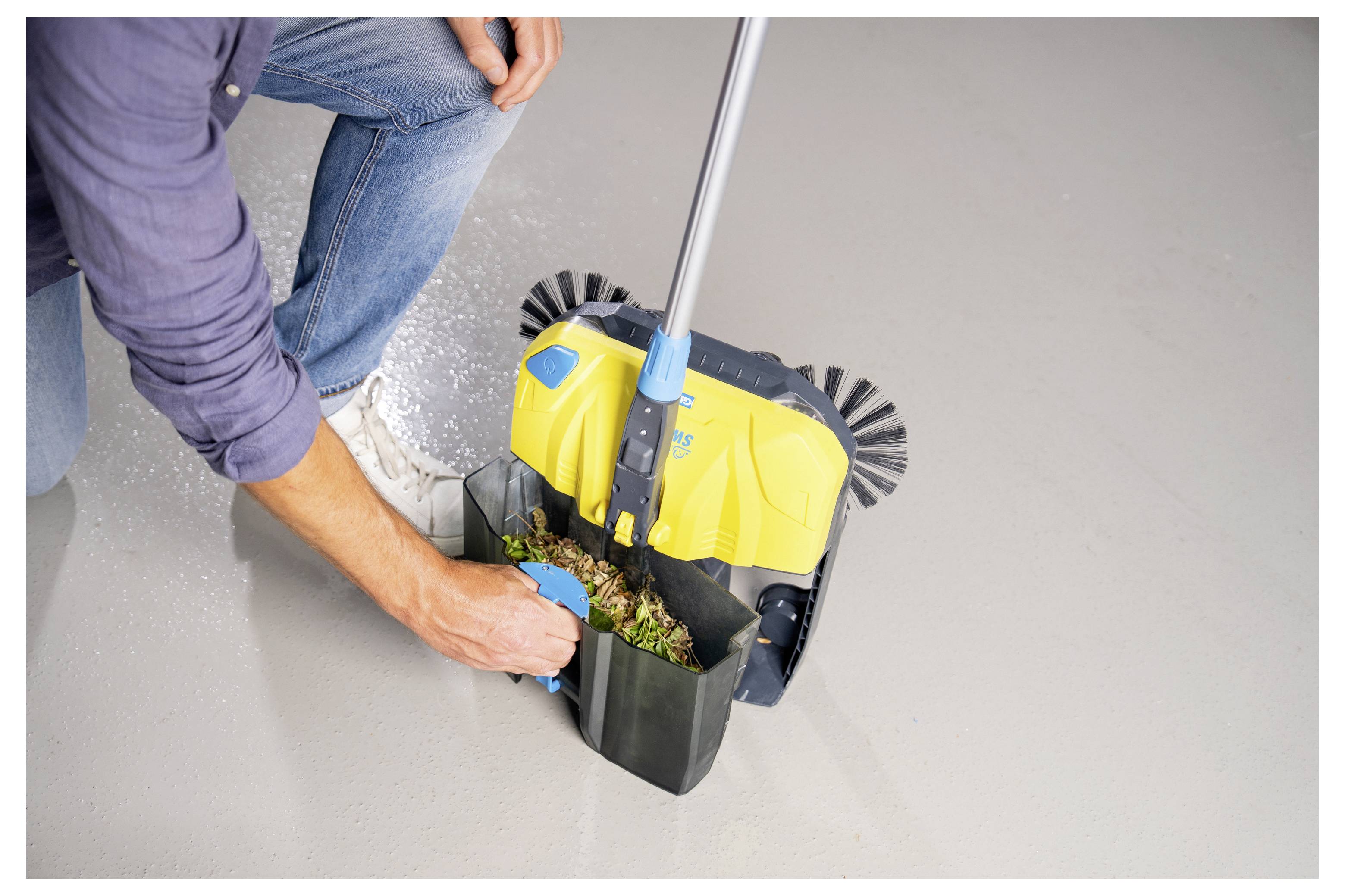 Eine Person hockt auf dem Boden und benutzt einen kleinen manuellen Kehrbesen, um Verschmutzungen zu sammeln, was die Funktionalität des Werkzeugs für Reinigungszwecke zeigt.