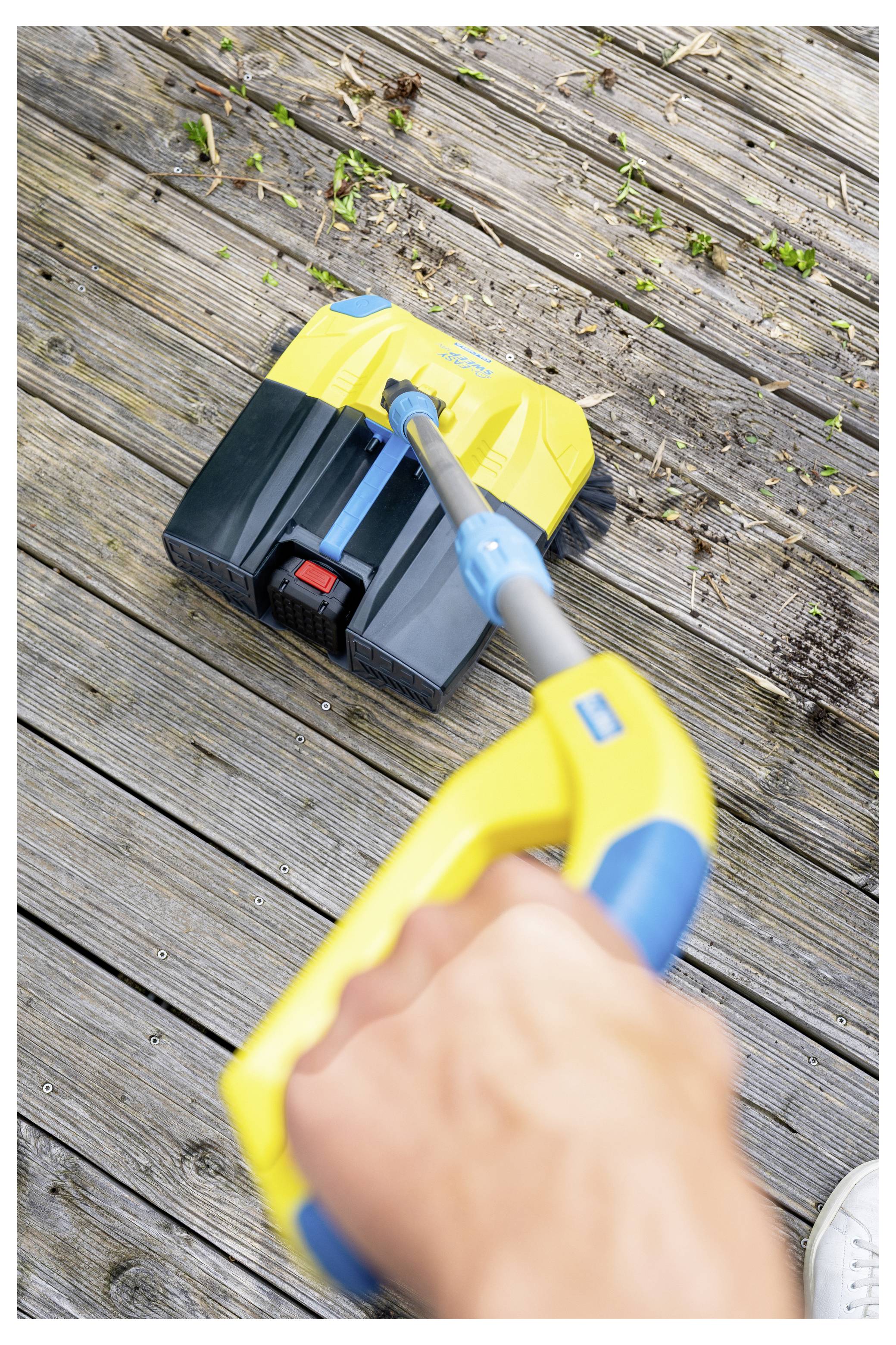 Eine Hand, die ein gelbes und blaues Reinigungsgerät hält, schrubbt ein Holzdeck, mit kleinen Trümmern, die herum verstreut sind.