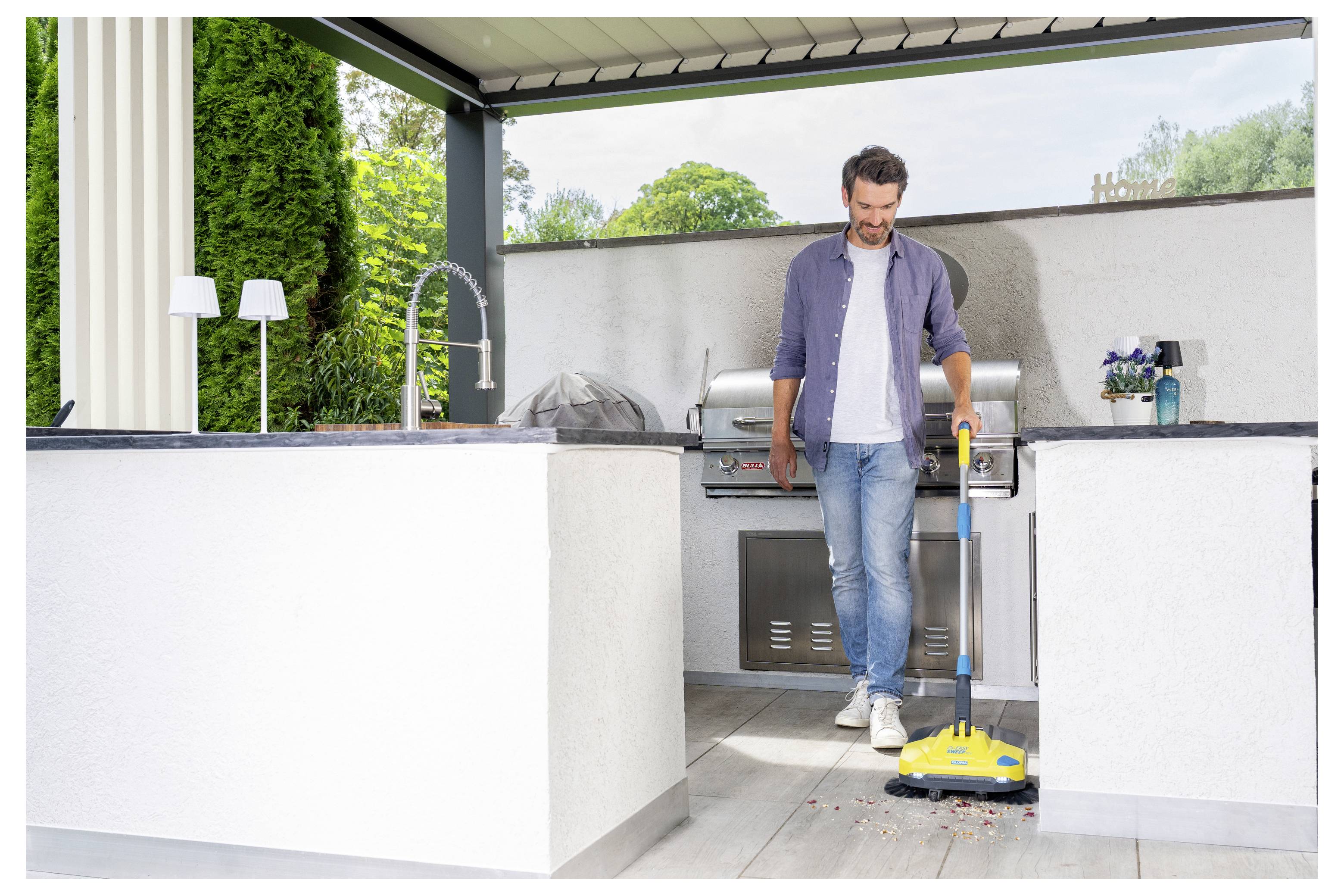 Ein Mann in legerer Kleidung kehrt die Terrasse einer Außenküche mit einem gelben Besen, während sich ein Grillgerät im Hintergrund befindet.