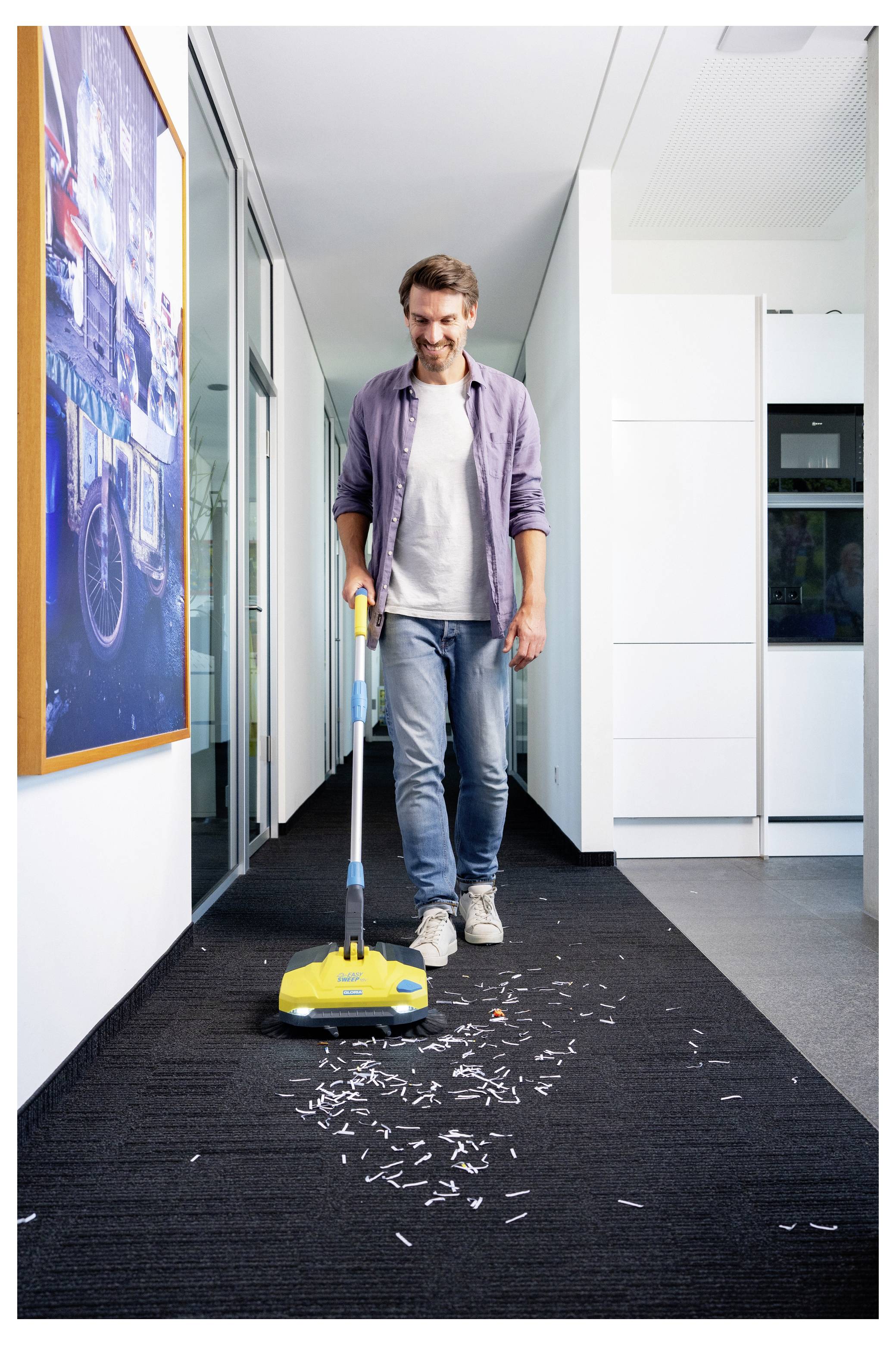 Eine Person lächelt und saugt Papierschnipsel mit einem Staubsauger aus einem Teppichboden in einem Büroflur, während moderne Kunst an der Wand hängt und die Umgebung sauber und hell ist.