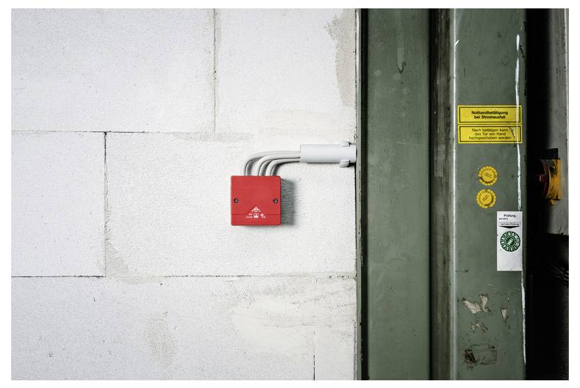 Rote Notfallalarmbox an einer weißen Wand neben einer grünen Industrietür mit Warnetiketten.