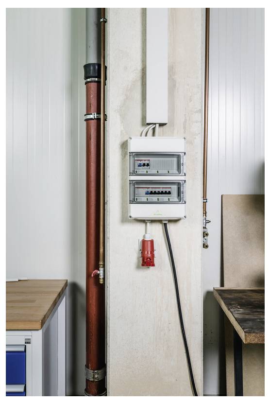 Elektrisches Schaltpanel und rotes Rohr auf einer unvollendeten Innenwand zwischen Werkbänken installiert, mit einem Kabel, das zu einer roten Steckdosendose führt.
