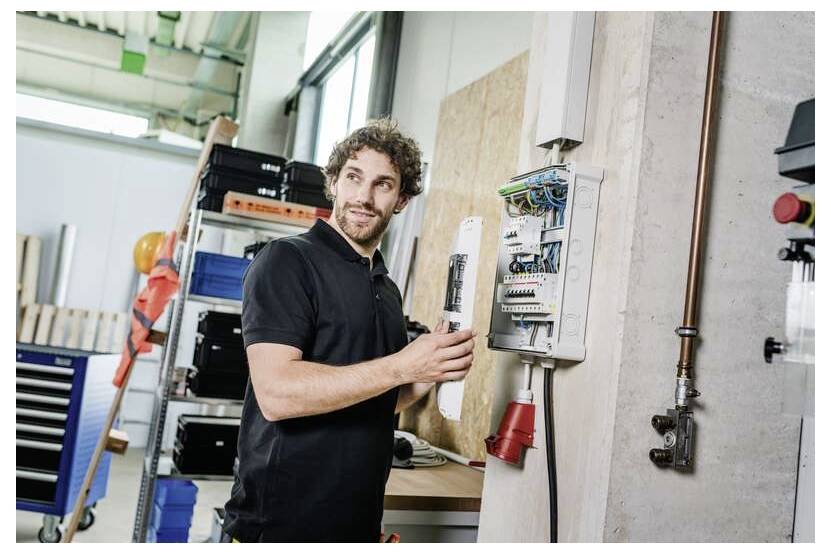 Techniker, der in einer Werkstatt ein elektrisches Schaltpult inspiziert, hält die Tür offen in der Nähe von Werkzeugen und Lagerregalen.