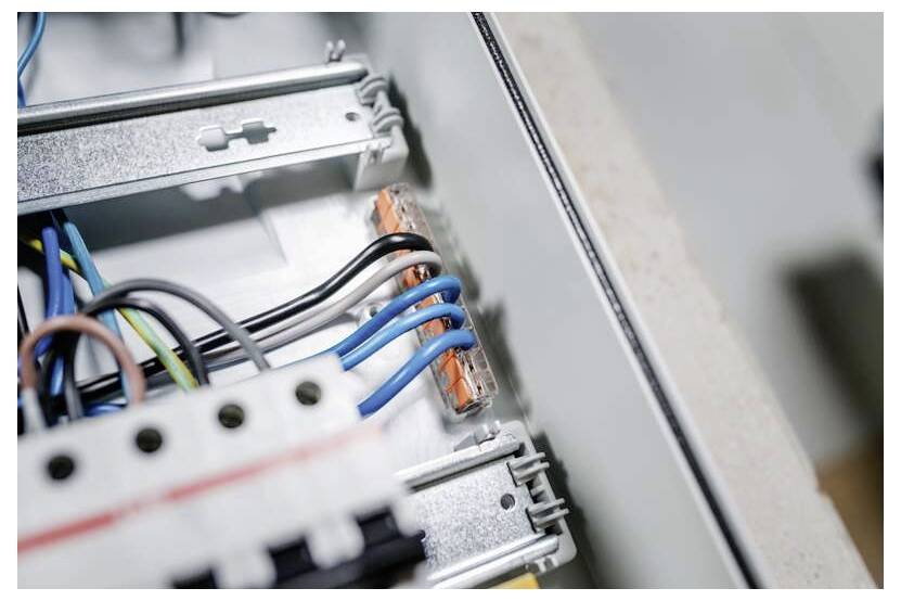 Nahaufnahme der elektrischen Verkabelung in einem Schaltschrank mit blauen, schwarzen und grünen Drähten, die an Metallklemmen angeschlossen sind.