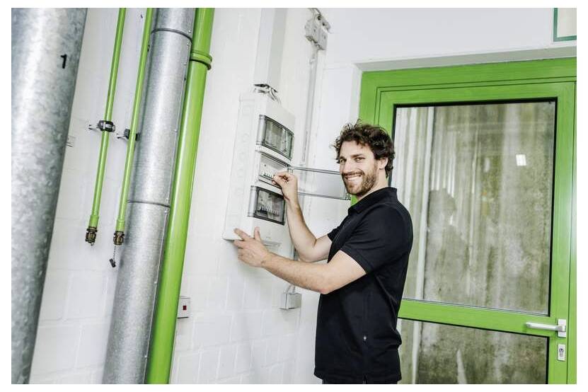 Techniker passt ein wandmontiertes Bedienfeld neben leuchtend grünen Rohren und einer Tür in einem Industriegebäude an.