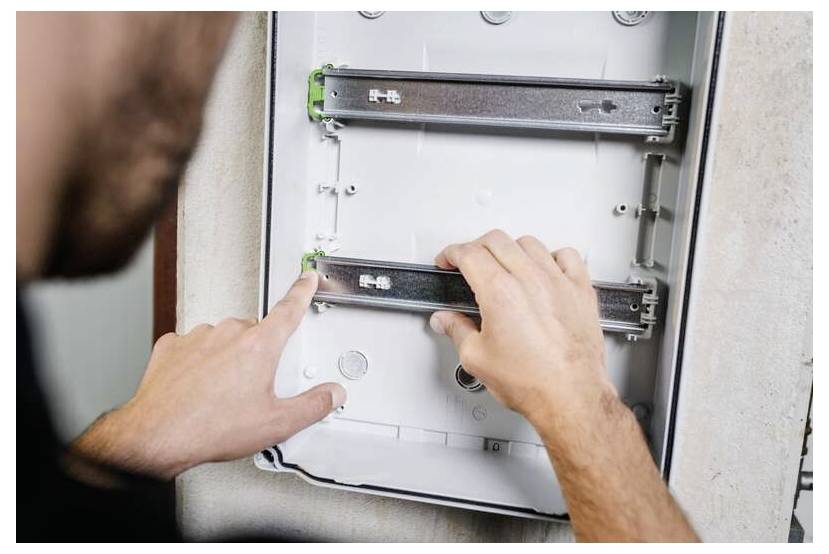 Person installiert ein wandmontiertes Elektroschaltfeld und stellt die verschiebbaren Metallschienen im offenen Kasten ein.