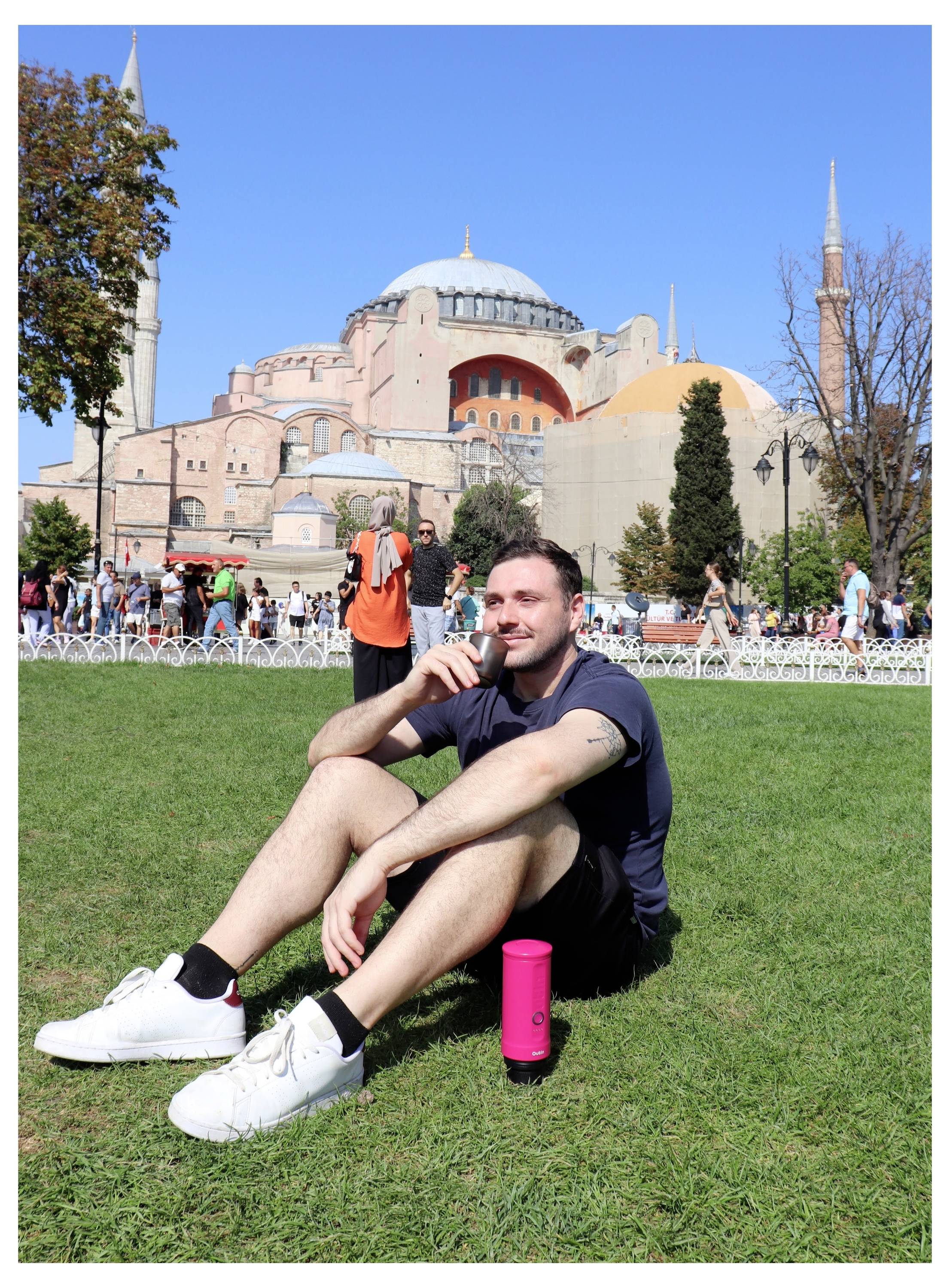 Eine Person sitzt auf Gras, lächelt, mit einem historischen Gebäude und einer Menschenmenge im Hintergrund unter einem klaren blauen Himmel.
