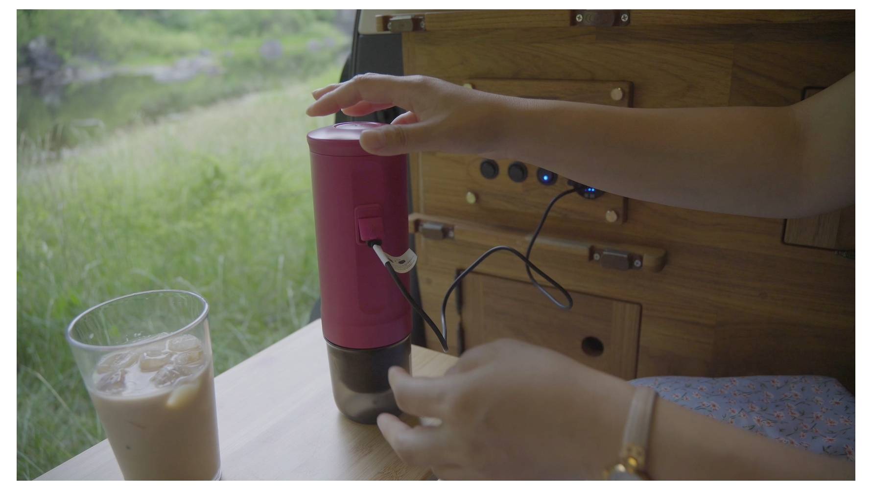 Eine Person benutzt eine tragbare Kaffeemühle in einem hölzernen Wohnmobil, neben einem Glas Eiskaffee auf einem Tisch, mit Blick auf eine grüne Wiese draußen.