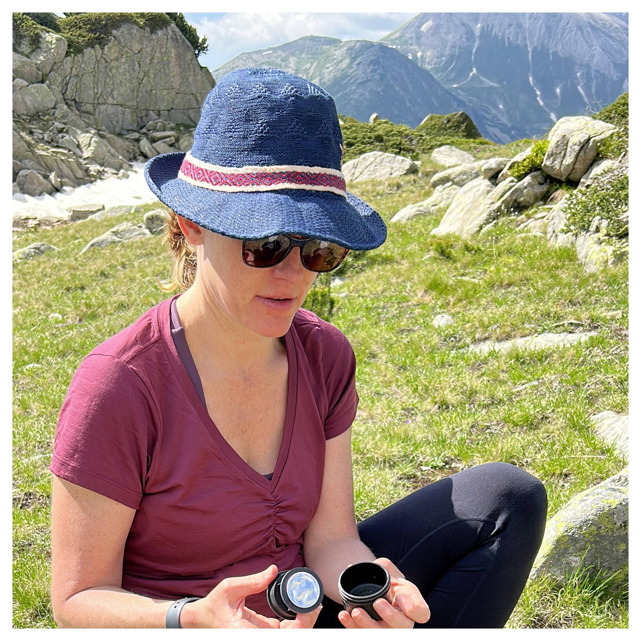 Eine Person mit einem blauen Hut und einer Sonnenbrille sitzt auf Gras mit Bergen im Hintergrund und untersucht einen schwarzen Gegenstand in ihrer Hand.