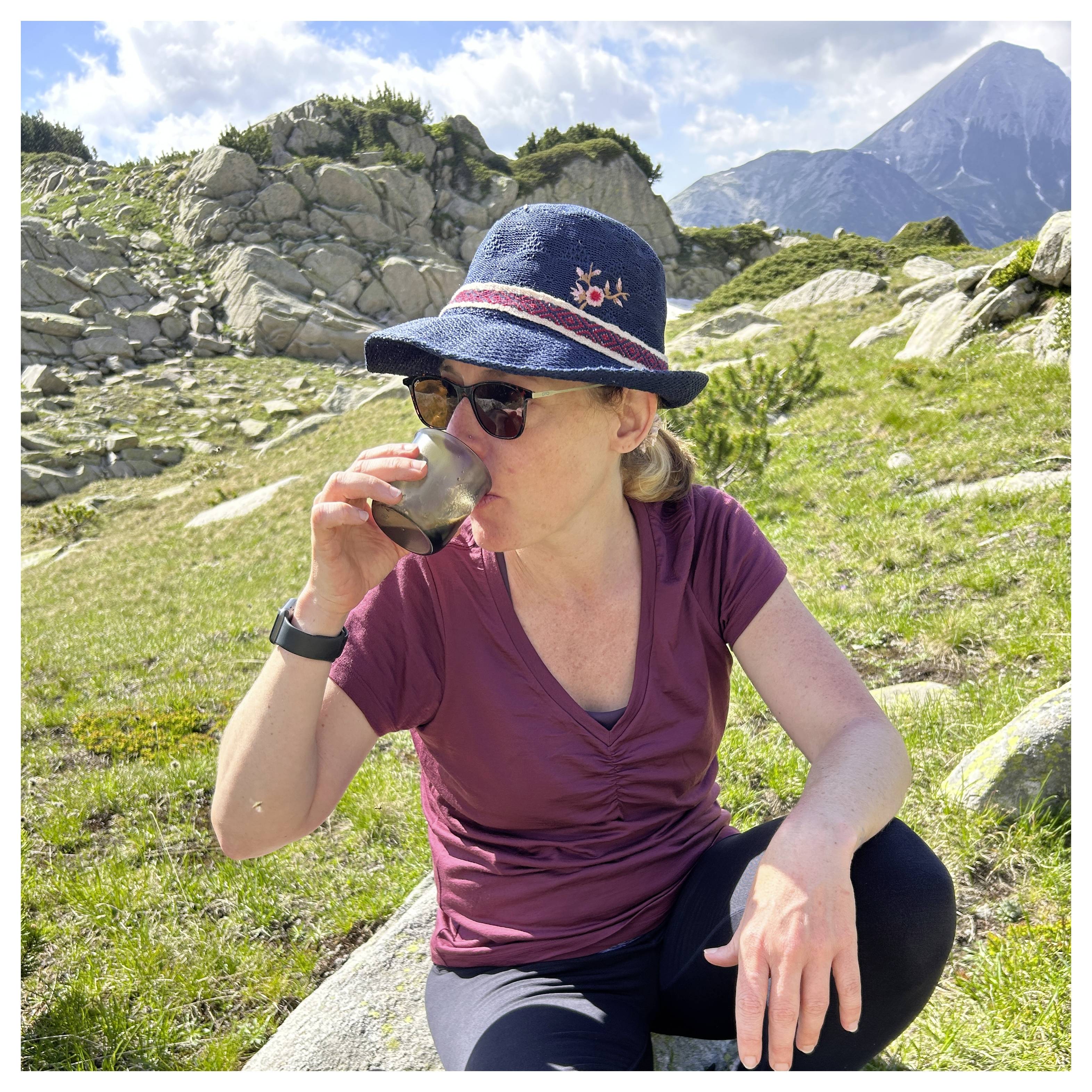 Frau mit Hut sitzt auf Gras und trinkt aus einer Tasse, mit felsiger Landschaft und Berg im Hintergrund.