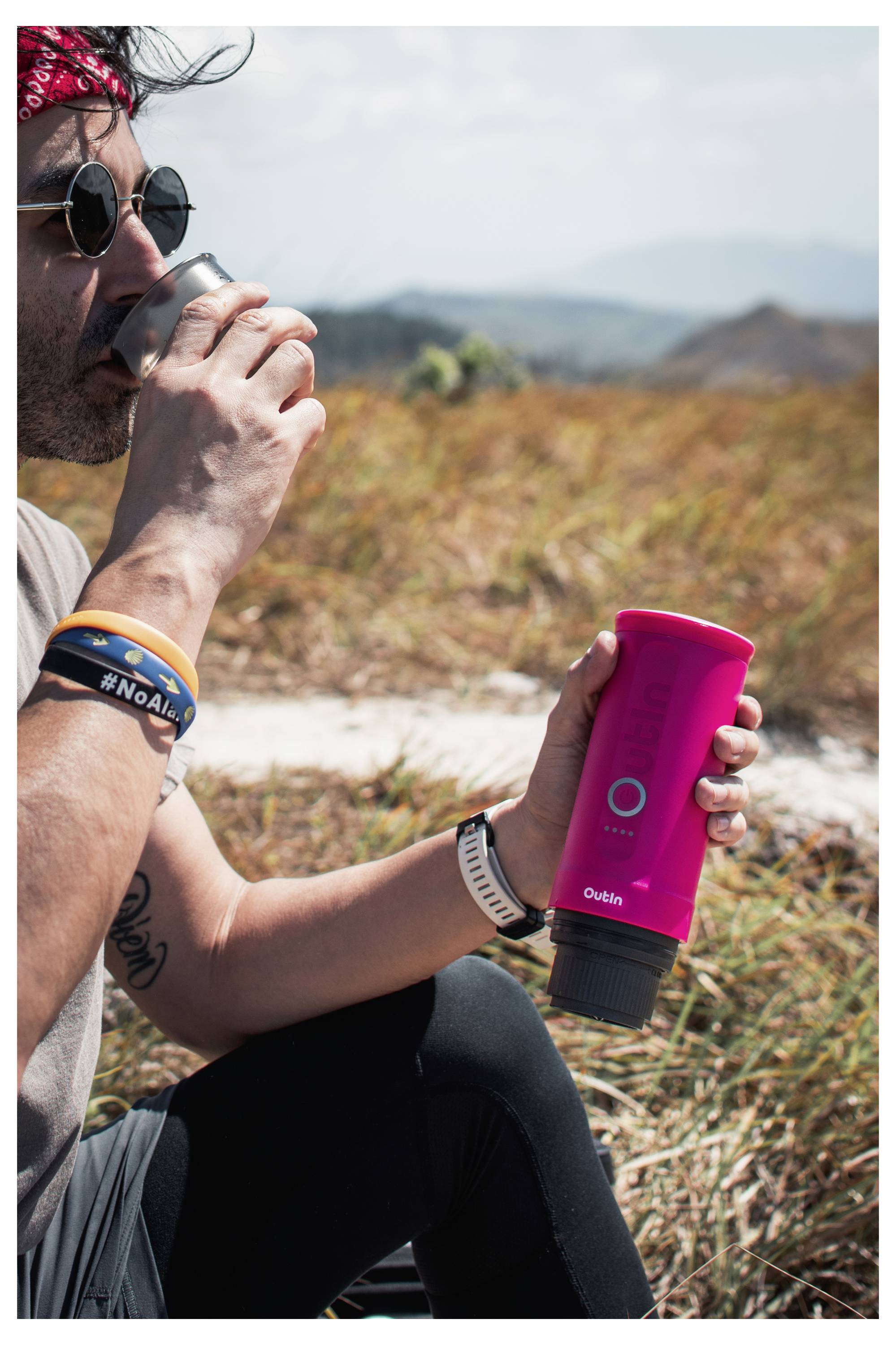 Eine Person im Freien auf einem Feld hält eine leuchtend rosa tragbare Kaffeemaschine mit der Marke 