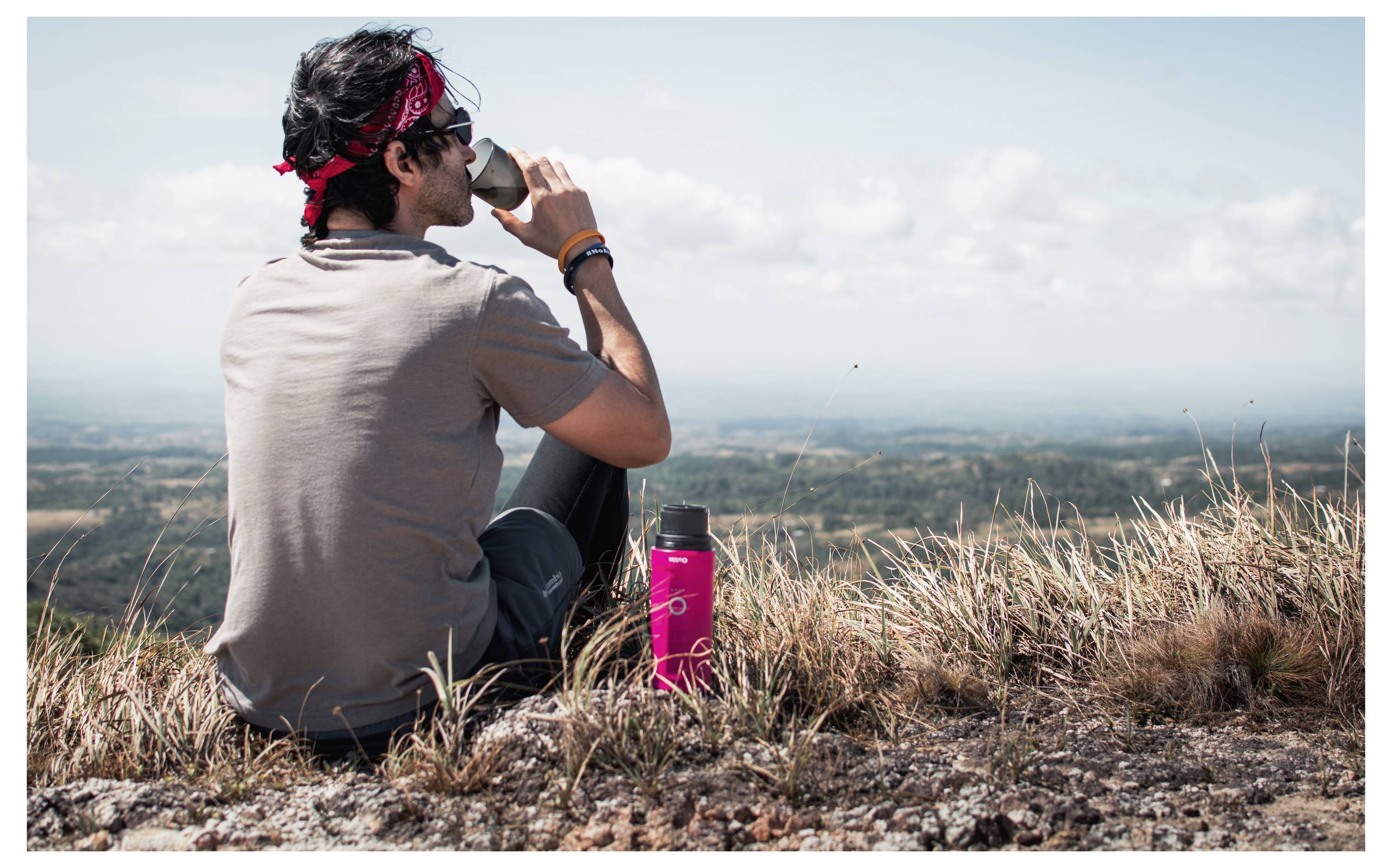 Eine Person sitzt auf Gras oben auf einem Hügel und trinkt aus einer Tasse. Eine rote Wasserflasche liegt in der Nähe. Der Hintergrund zeigt eine weite, malerische Landschaft.