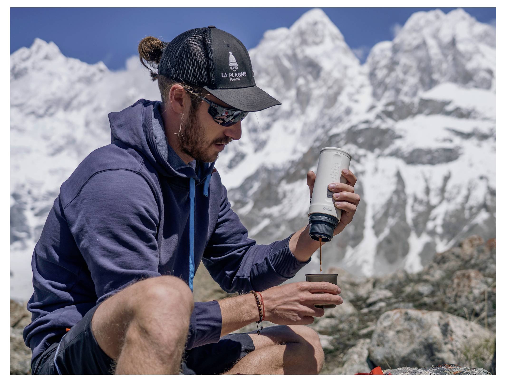 Ein Mann in Outdoor-Ausrüstung und Sonnenbrille bereitet ein heißes Getränk mit einer tragbaren Kaffeemaschine vor einem Hintergrund von schneebedeckten Bergen zu.
