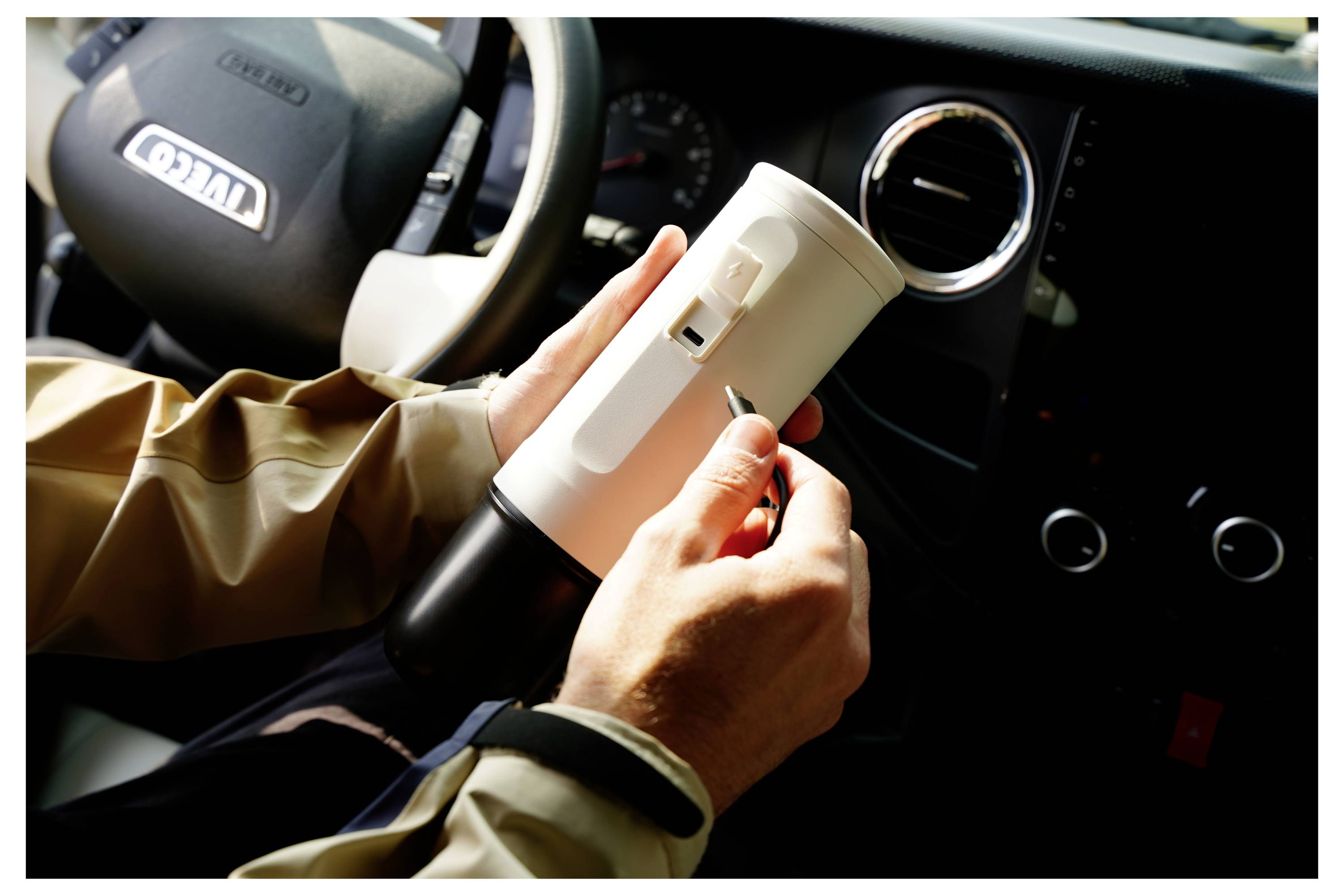 Eine Person in einem Fahrzeug hält einen weißen zylindrischen Gegenstand, der einem kleinen Haushaltsgerät ähnelt, und verstellt einen Schalter an seiner Seite. Das Armaturenbrett ist sichtbar.