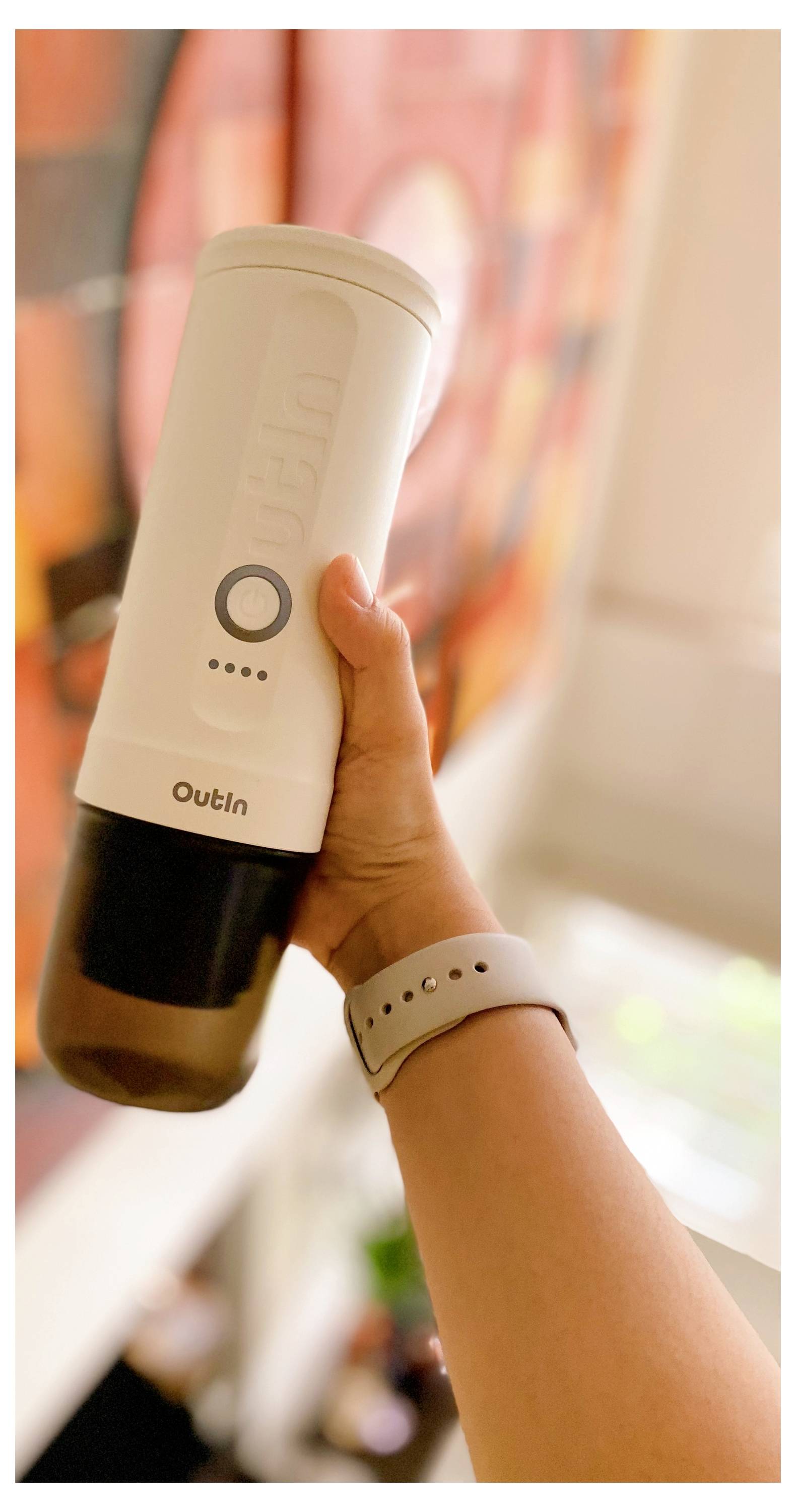 Eine Person hält eine tragbare elektrische Kaffeemühle in einem gut beleuchteten Raum, mit einem unscharfen bunten Gemälde im Hintergrund.