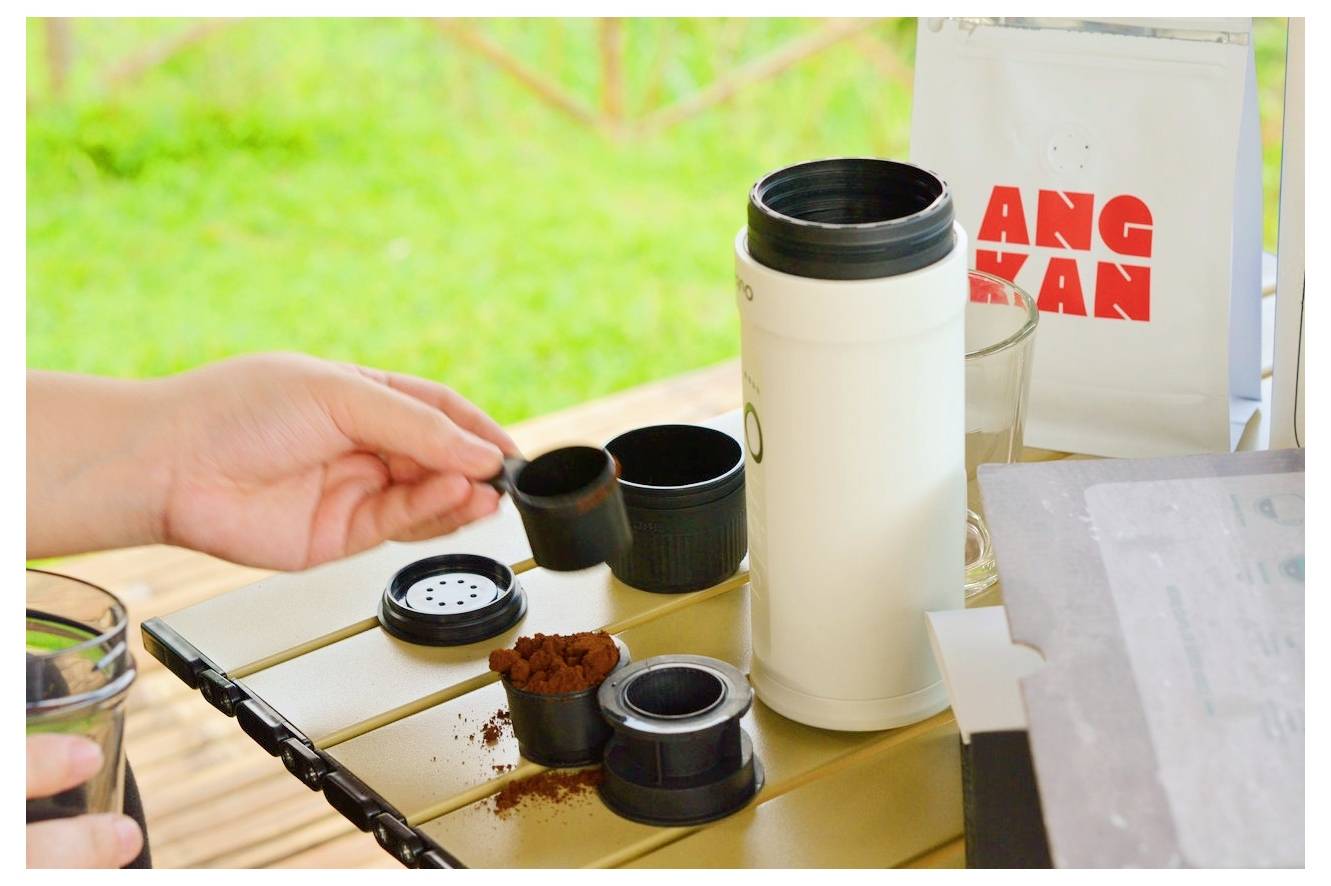 Eine Person bereitet Kaffee im Freien mit einer tragbaren Kaffeemaschine zu. Gemahlener Kaffee wird in einem Filter neben der Kaffeemaschine platziert.