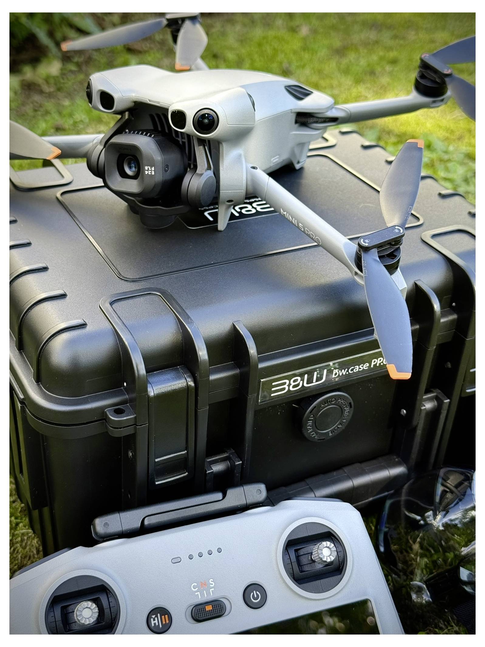 Eine Drohne mit Kamera sitzt auf einem schwarzen Schutzkoffer, während die Fernbedienung in der Nähe auf dem Gras liegt.