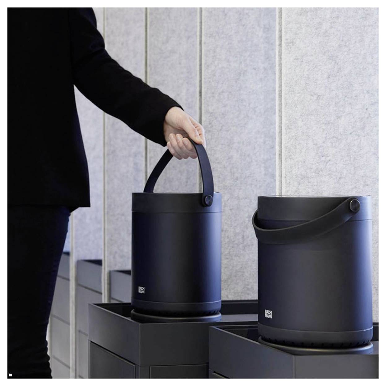 Eine Person in dunkler Kleidung stellt einen eleganten, schwarzen, zylindrischen Behälter mit Griff auf eine graue Oberfläche, was auf Tragbarkeit hindeutet.