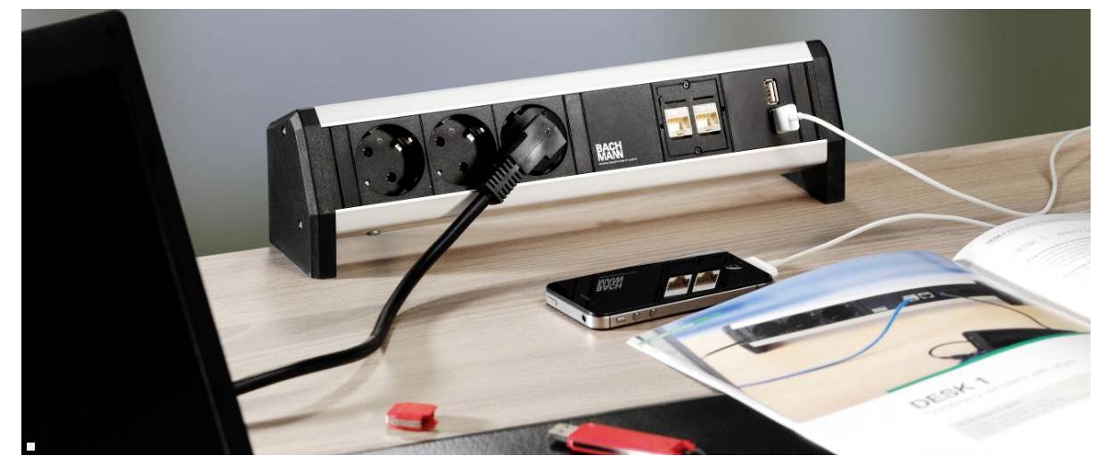 Mehrfachstecker mit mehreren Steckdosen und USB-Anschlüssen auf einem Schreibtisch, die ein Telefonladegerät anschließen. Bücher und ein Smartphone befinden sich in der Nähe.