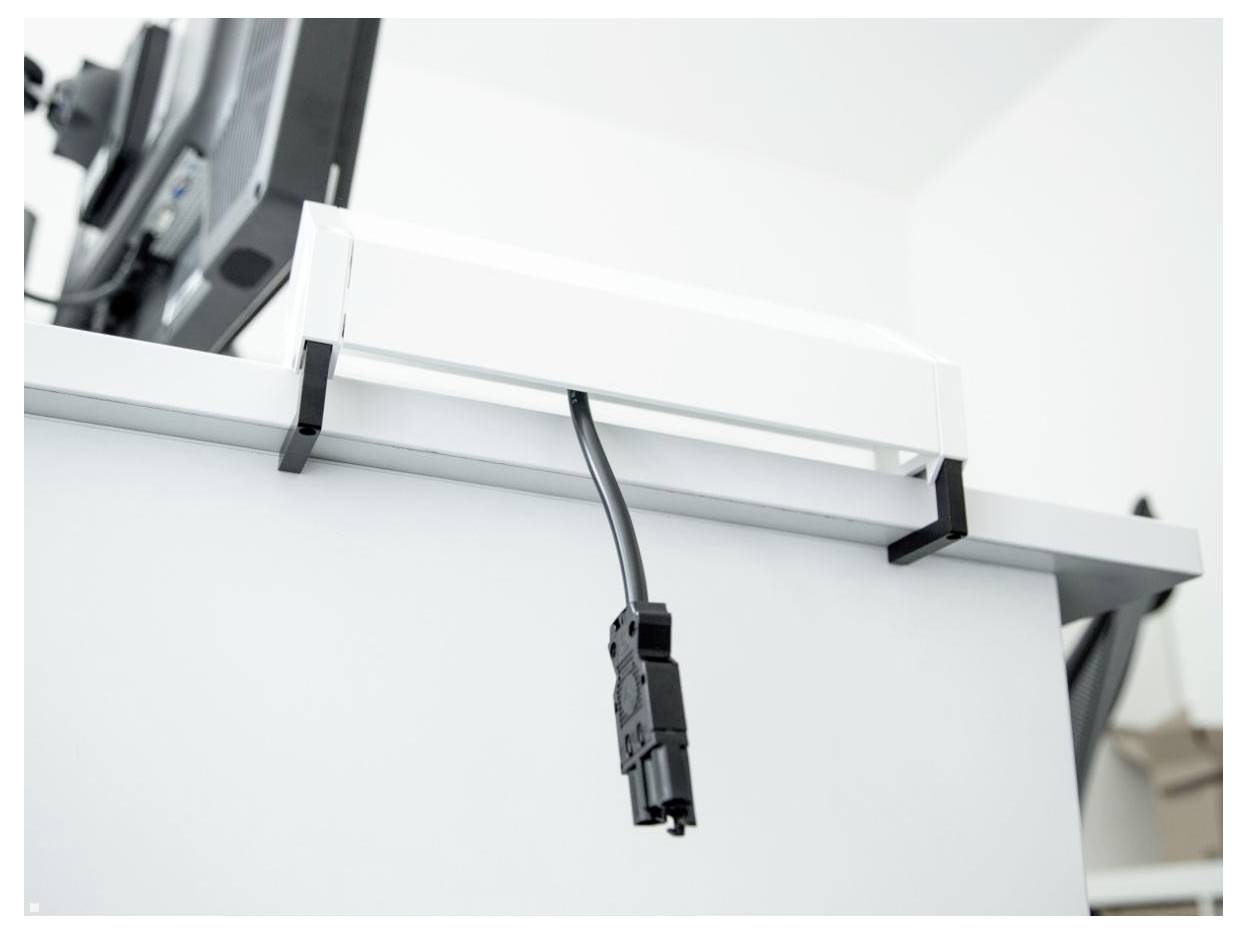 Ein schwarzes Kabel, das durch Clips gesichert ist, hängt von der Unterseite eines weißen Schreibtisches herab, neben einem Monitor auf einem Schwenkarm in einer modernen Büroumgebung.