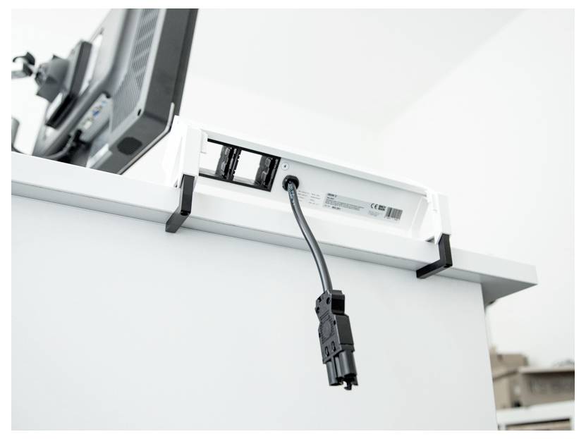 Ein eingestöpseltes Stromkabel hängt von einer Steckdosenleiste unter einem Bürotisch herab, auf dem ein Computermonitor steht, was auf eine unvollständige Einrichtung hindeutet.