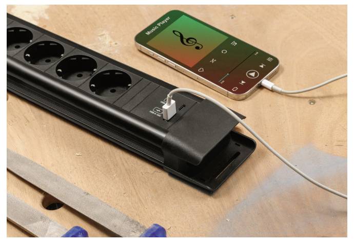 Ein Smartphone, das Musik abspielt, ist mit einem USB-Kabel an eine Steckdosenleiste angeschlossen und liegt auf einer Holzoberfläche neben einer Metallfeile.