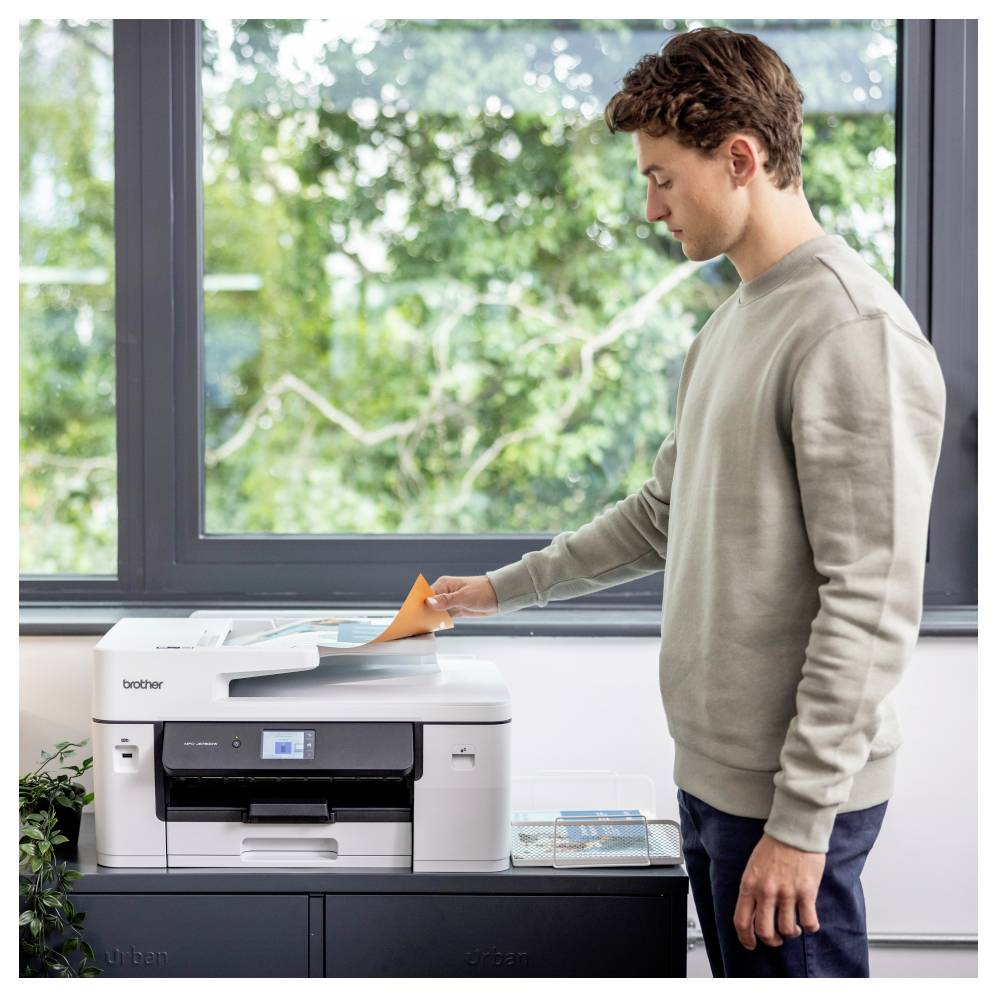 Ein Mann bedient einen Drucker und führt oranges Papier in den Scanner ein. Die Szene spielt sich in einem hellen Raum mit einem großen Fenster und Grünflächen außerhalb ab.