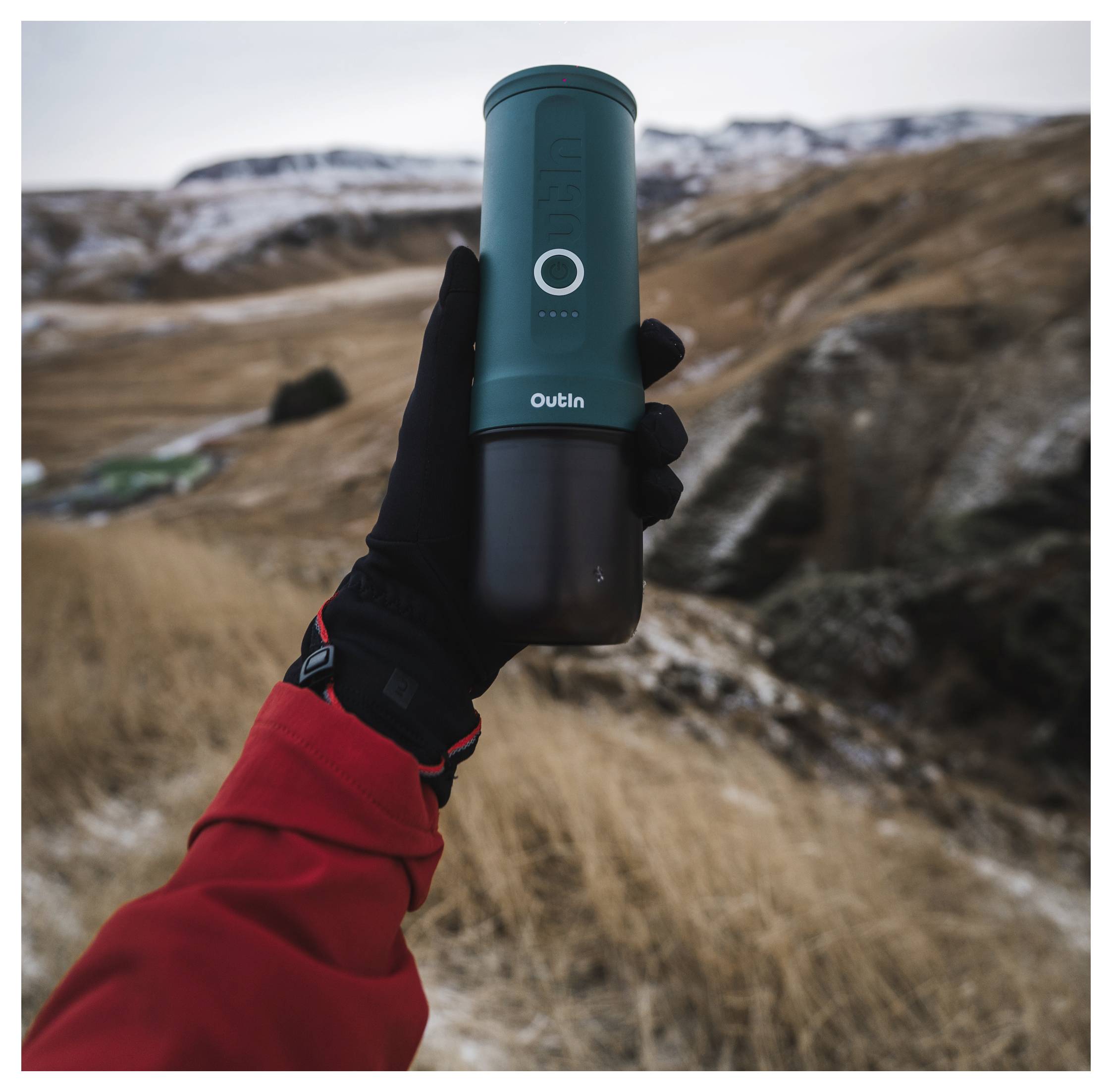 Eine Person in einer roten Jacke und schwarzem Handschuh hält eine tragbare Thermoskanne in einer bergigen Landschaft und symbolisiert Outdoor-Abenteuer.