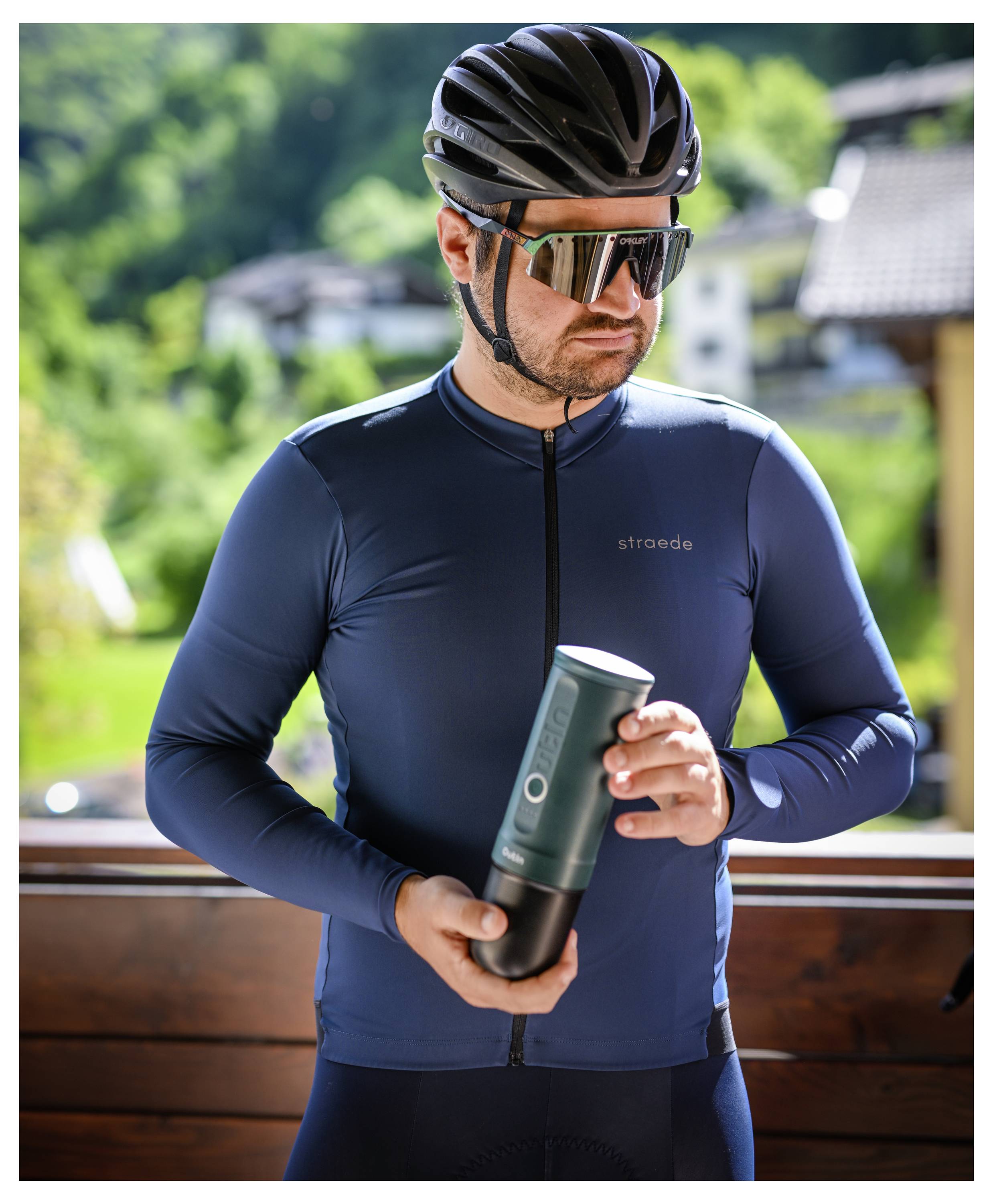Ein Radfahrer mit Sonnenbrille und Helm hält eine Trinkflasche zum Radfahren im Freien, mit Häusern und Grünflächen im Hintergrund.