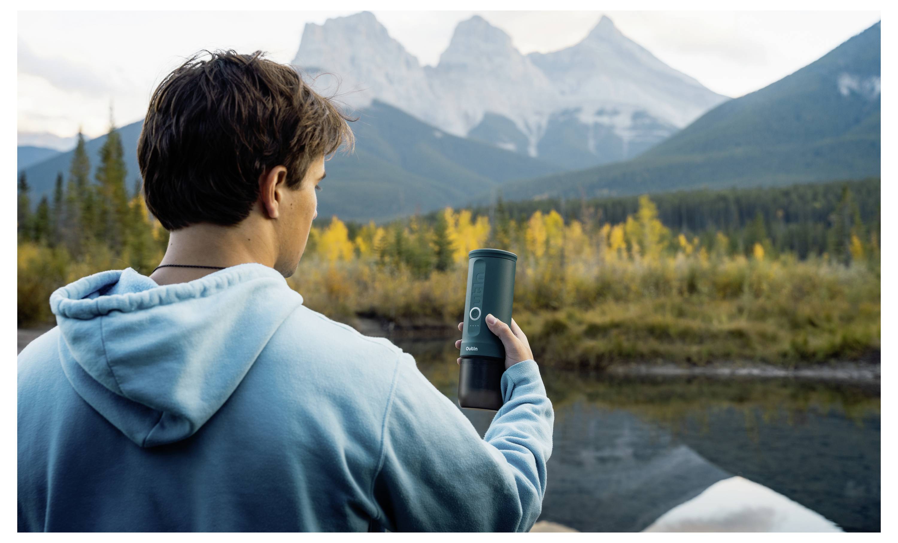 Eine Person in einem blauen Hoodie hält einen Thermobecher und blickt auf Bergspitzen, die sich über eine bewaldete Landschaft mit einem reflektierenden Gewässer erstrecken.