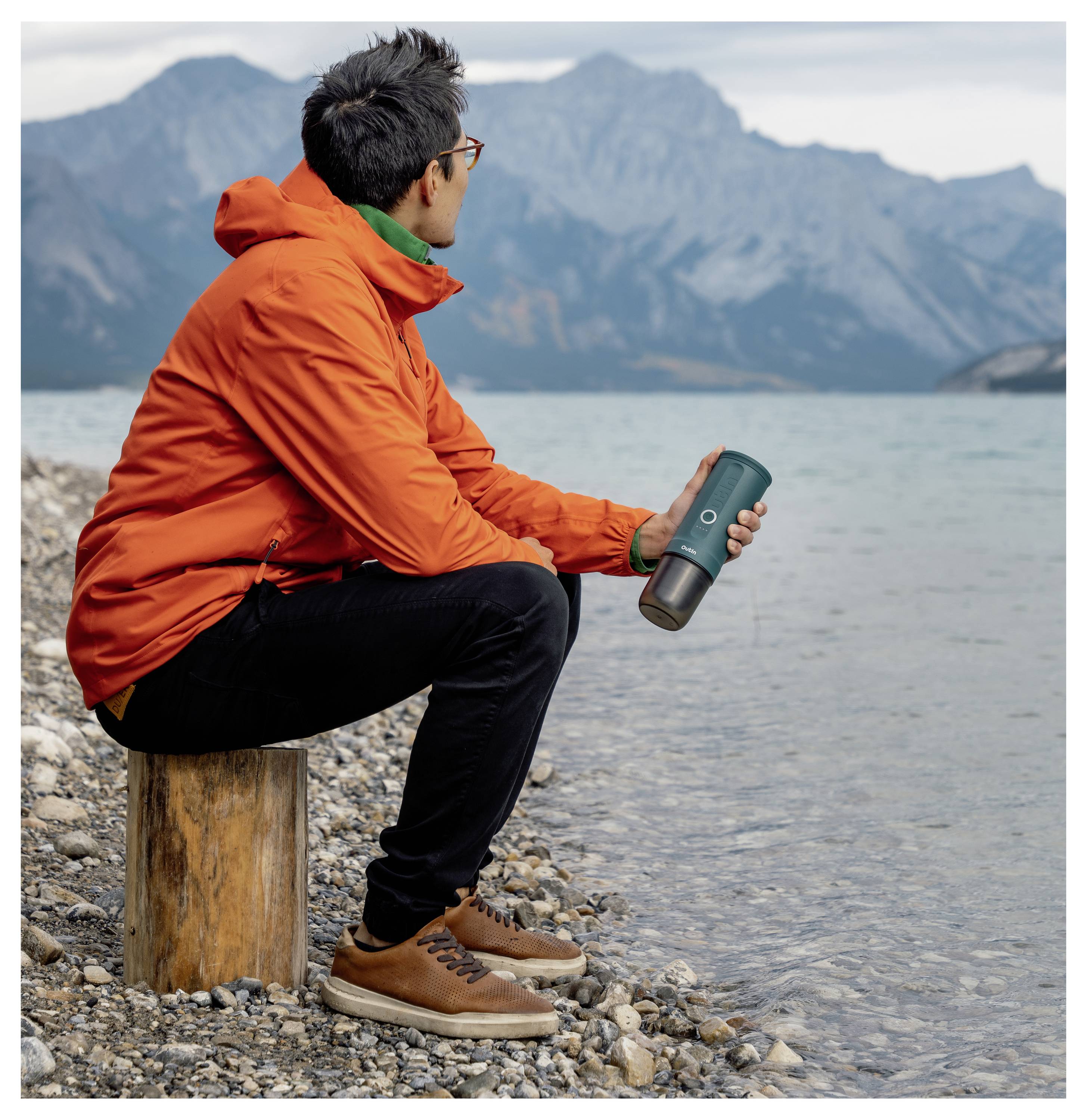 Eine Person in einer orangefarbenen Jacke sitzt auf einem Baumstamm am See und hält eine Thermoskanne. Im Hintergrund sind felsige Berge sichtbar.