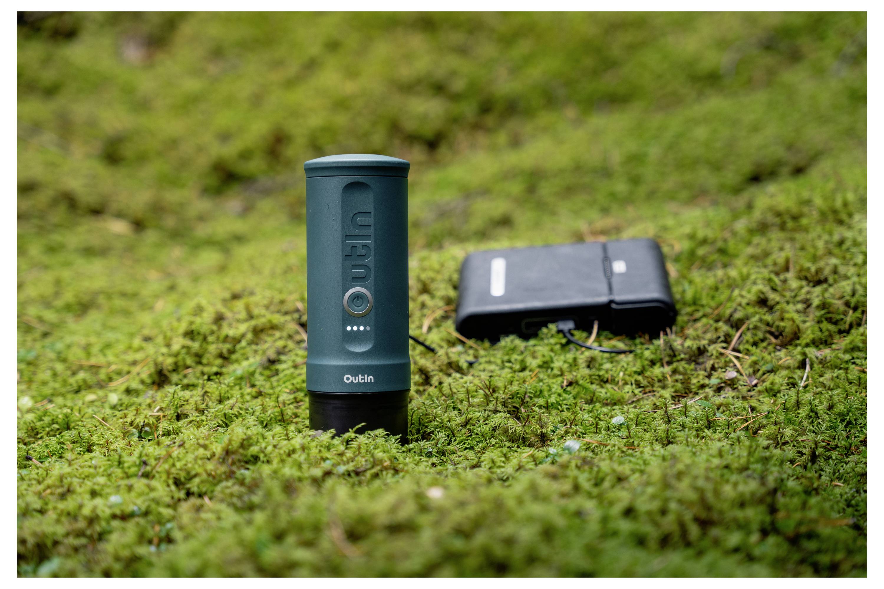 Eine tragbare Powerbank und ein kompakter Campingkocher sind auf grünem Moos platziert und deuten auf Outdoor-Nutzung und Komfort für Reisen hin.