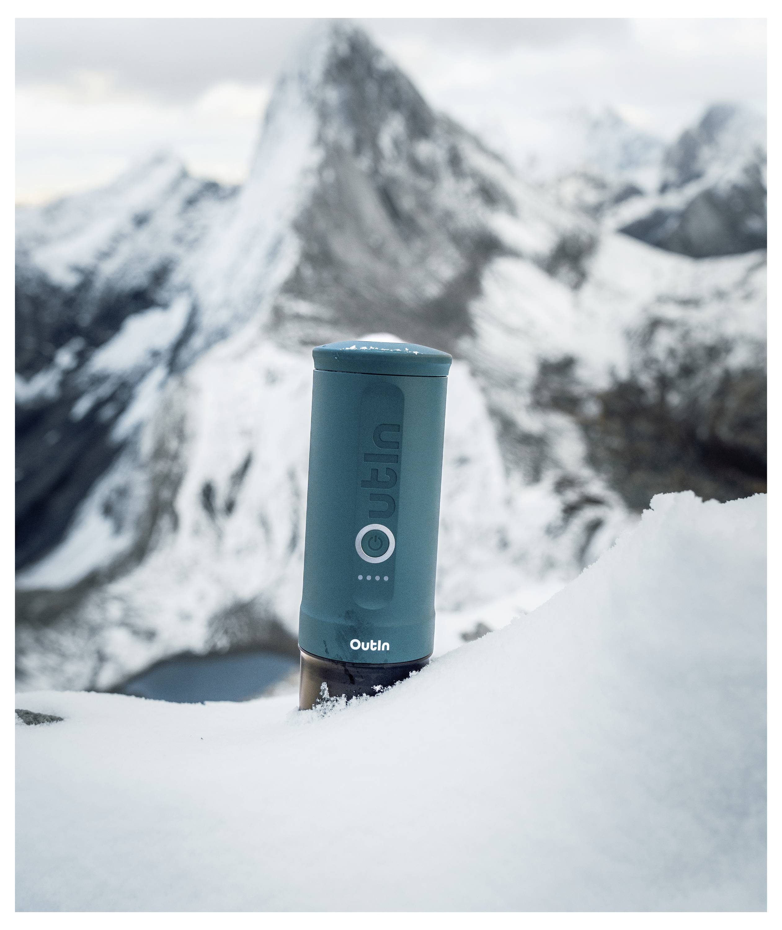 Eine blau gefärbte Outin-Markenthermosflasche ist teilweise im Schnee vergraben mit einem Hintergrund von schneebedeckten Bergen unter einem bewölkten Himmel.