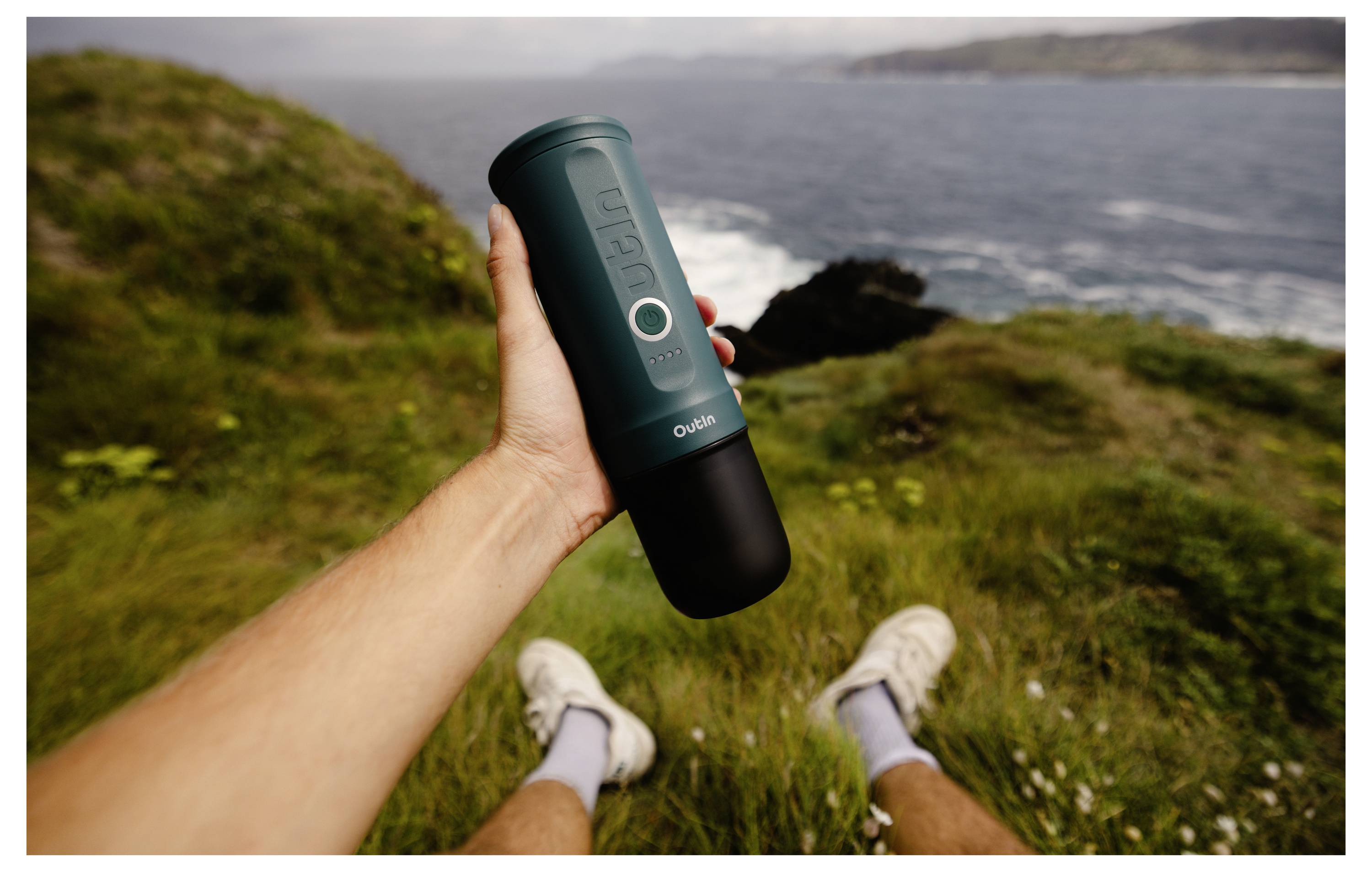 Eine Person hält eine tragbare Kaffeemaschine auf einer grasbewachsenen Klippe mit Blick auf den Ozean, mit einem fernen Ausblick auf Küstenlinien unter einem bewölkten Himmel.