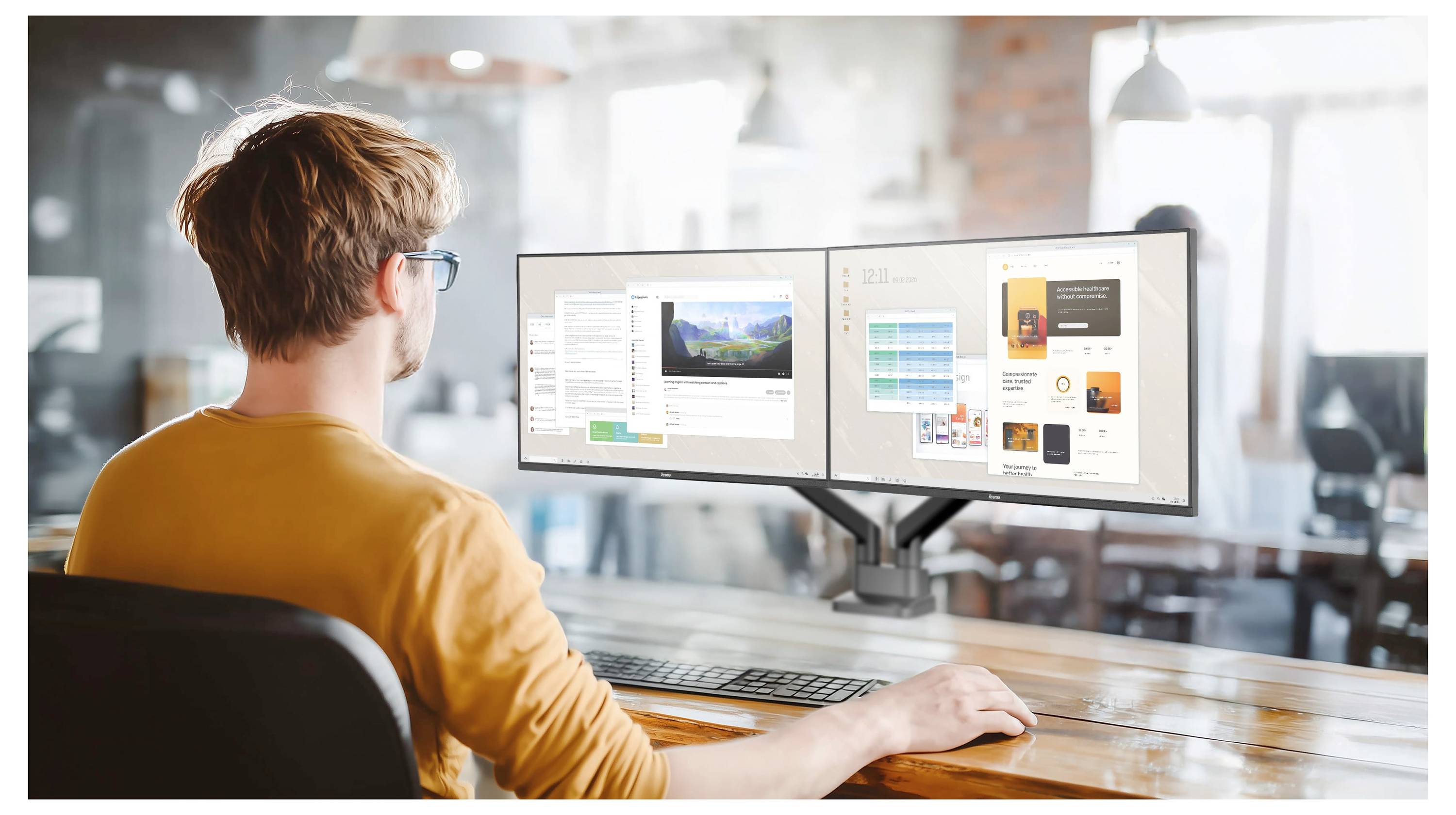 Eine Person, die an einem Holzschreibtisch mit zwei Computermonitoren in einem hellen, modernen Büro mit mehreren Fenstern und anderen Arbeitsplätzen im Hintergrund arbeitet.