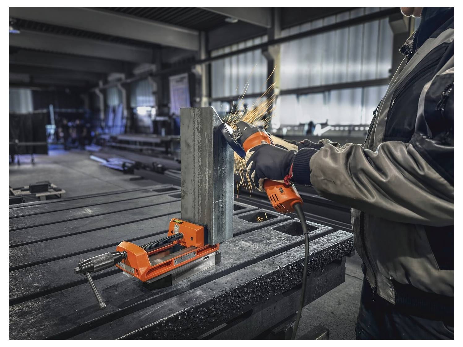Eine Person benutzt einen Winkelschleifer auf einem in einem Schraubstock befestigten Metallblock und erzeugt dabei Funken in einer industriellen Werkstattumgebung.