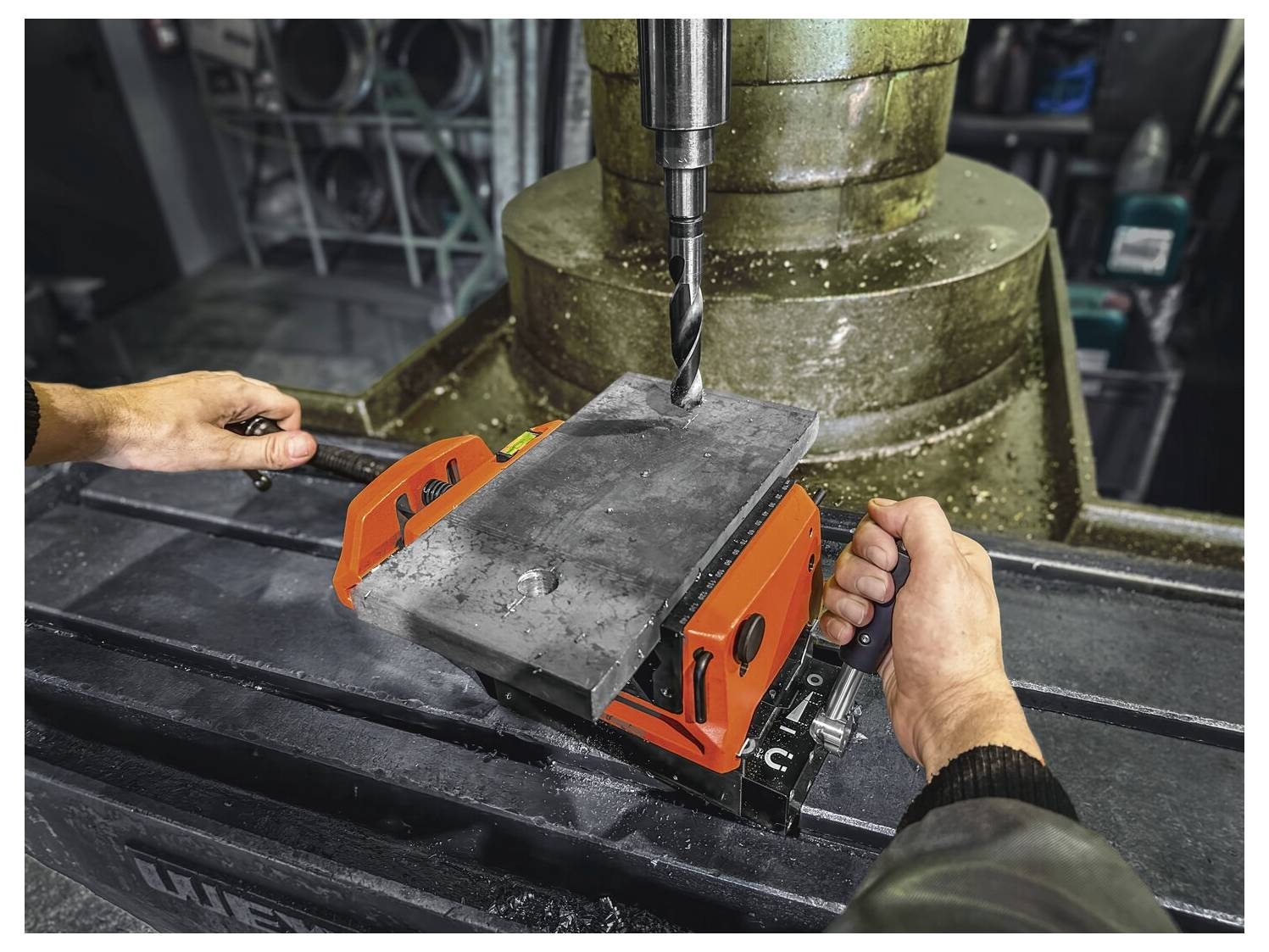 Zwei Personen bedienen eine Bohrmaschine und konzentrieren sich auf die Ausrichtung eines Metallstücks unter dem Bohrmeißel, wobei sie Teamfähigkeit und Präzision in einer Werkstattsumgebung demonstrieren.