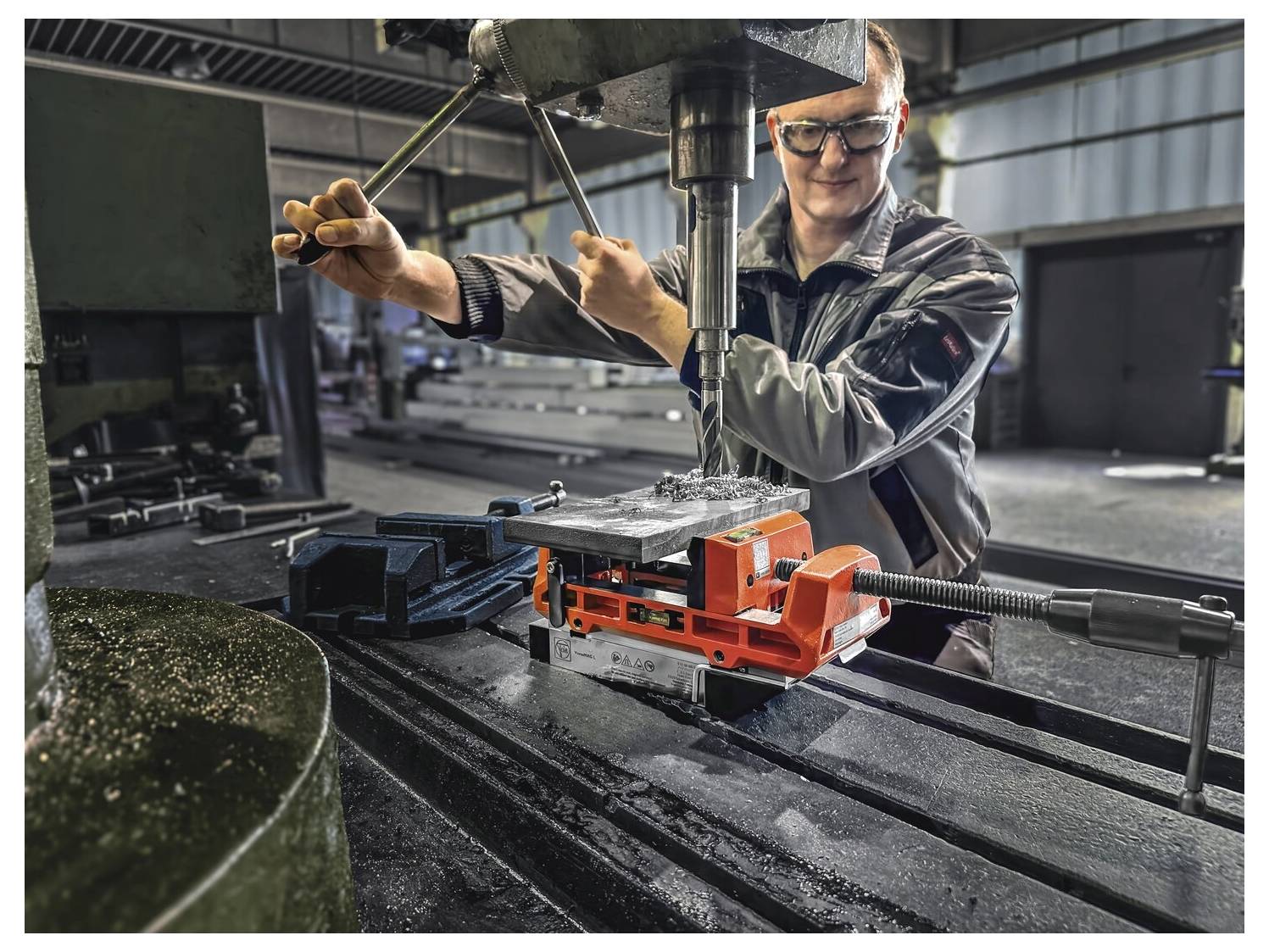 Eine Person bedient eine Industriebohrmaschine in einer Werkstatt und verwendet einen Metallschraubstock, um ein Werkstück zum Bohren zu sichern.