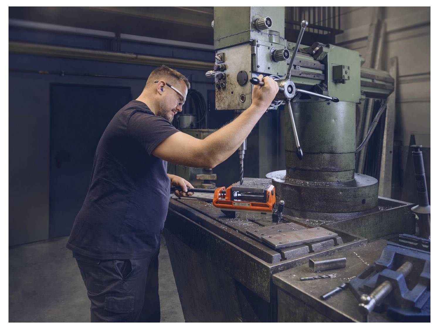 Eine Person bedient eine Fräsmaschine in einer Werkstatt und stellt ein Bauteil auf dem Tisch ein. Verschiedene Werkzeuge und Ausrüstungen sind in der Nähe sichtbar.