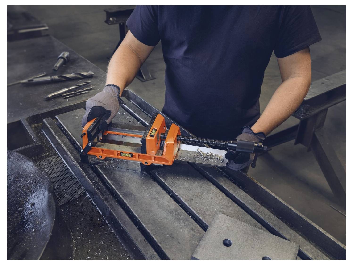 Eine Person verwendet eine orangefarbene Metallzwinge auf einer Werkbank mit Werkzeugen in der Nähe, trägt Handschuhe und ein dunkles Hemd in einer Industrieumgebung.
