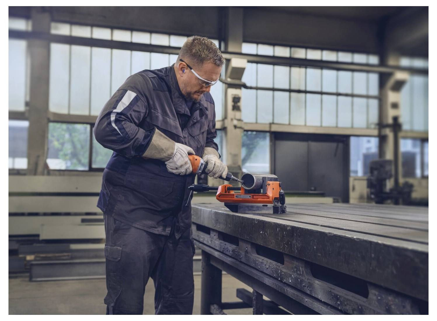 Ein Arbeiter in Schutzausrüstung schneidet ein Metallrohr auf einer industriellen Bandsäge an einer Werkbank in einer großen Werkstatt.