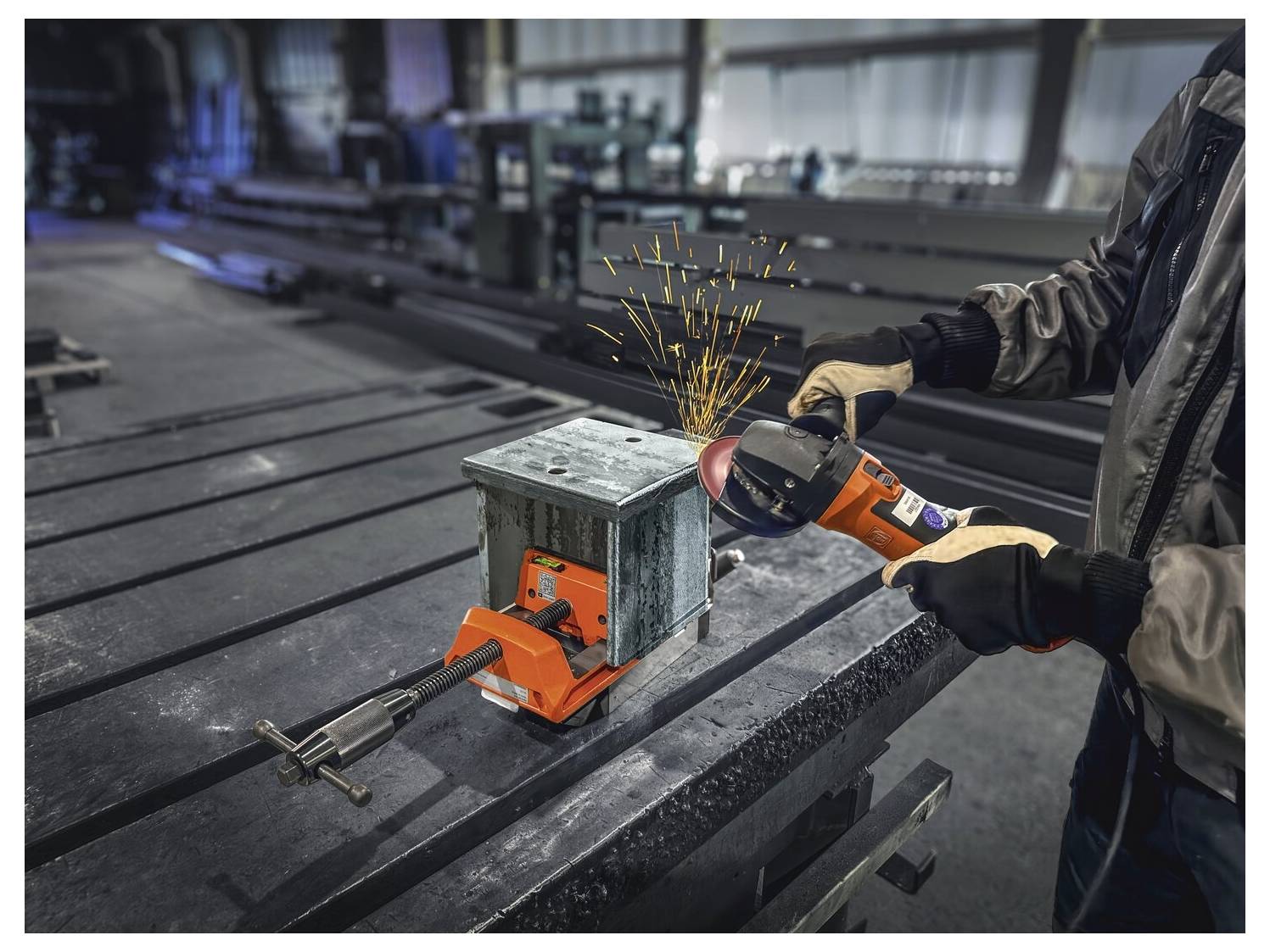 Ein Arbeiter schleifen eine Metallkiste, die auf einem Tisch eingespannt ist, und erzeugt dabei Funken. Die Werkstattumgebung umfasst Industrieausrüstung und Metalloberflächen.