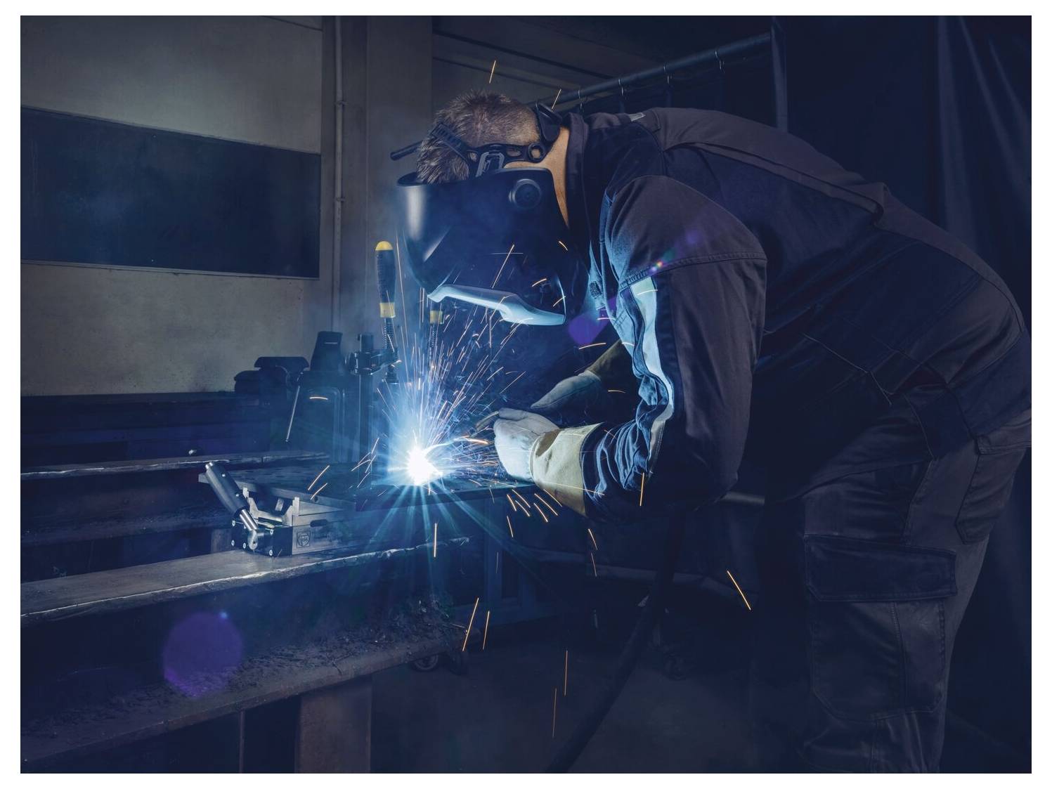 Eine Person in Schutzausrüstung schweißt Metall und erzeugt dabei helle Funken in einer schwach beleuchteten Werkstatt.