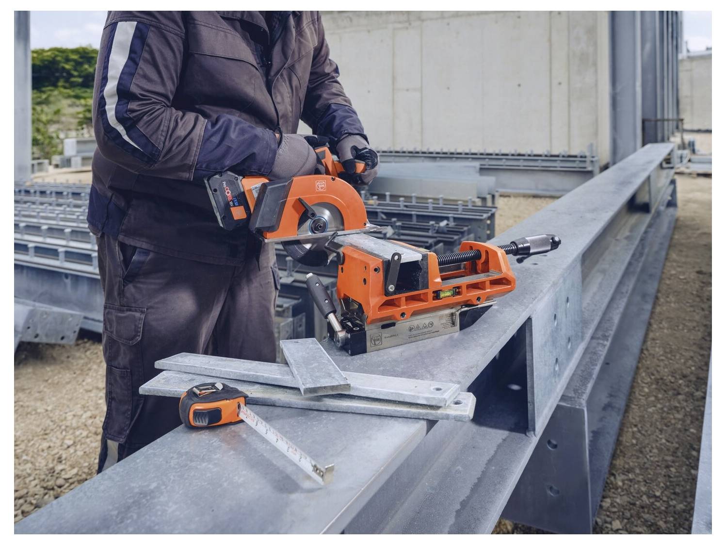 Eine Person benutzt eine orangefarbene Metallsäge an einem großen Stahlträger auf einer Baustelle. Ein Messstab und Stahlwinkel befinden sich in der Nähe.