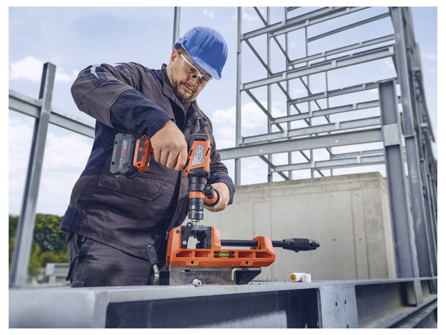 Ein Bauarbeiter in Schutzausrüstung bohrt mit einer Bohrmaschine in Metallträger auf einer Baustelle. Ein teilweise errichtetes Stahlgerüst ist sichtbar.